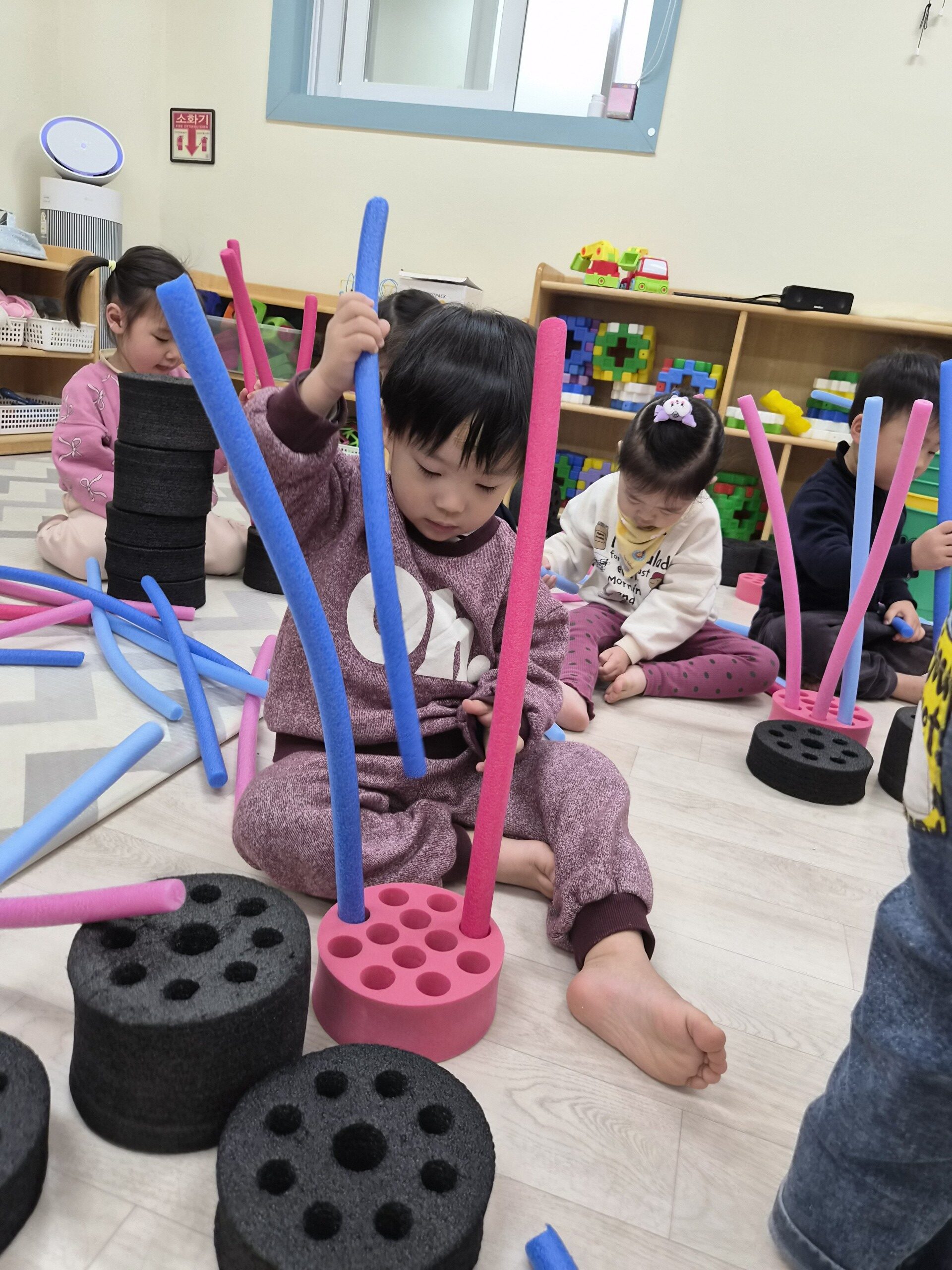 지호가 자주색 상하의를 입고 맨발로 앉아 분홍색 구멍 블록에 길고 유연한 파란색과 분홍색 스틱을 끼워 넣으며 집중하고 있다. 주변에는 다른 아이들도 같은 놀이를 하고 있다.
