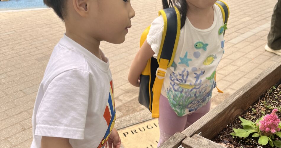 야외 화단에서 여자아이와 함께 허리를 굽혀 작은 식물들을 유심히 들여다보는 지호의 모습. 지호는 흰색 티셔츠와 줄무늬 반바지를 입고 있고, 여자아이는 왕관을 쓰고 작은 노란색 배낭을 메고 있다.