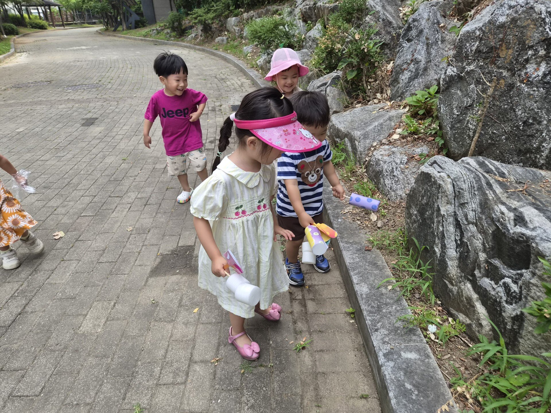 숲길 옆 포장된 길을 따라 여러 명의 아이들이 걸어가고 있다. 지호는 분홍색 티셔츠를 입고 즐거운 표정으로 다른 아이들과 함께 옆쪽의 돌멩이와 풀을 바라보고 있다. 한 여자아이는 분홍색 선캡을 쓰고 있다.