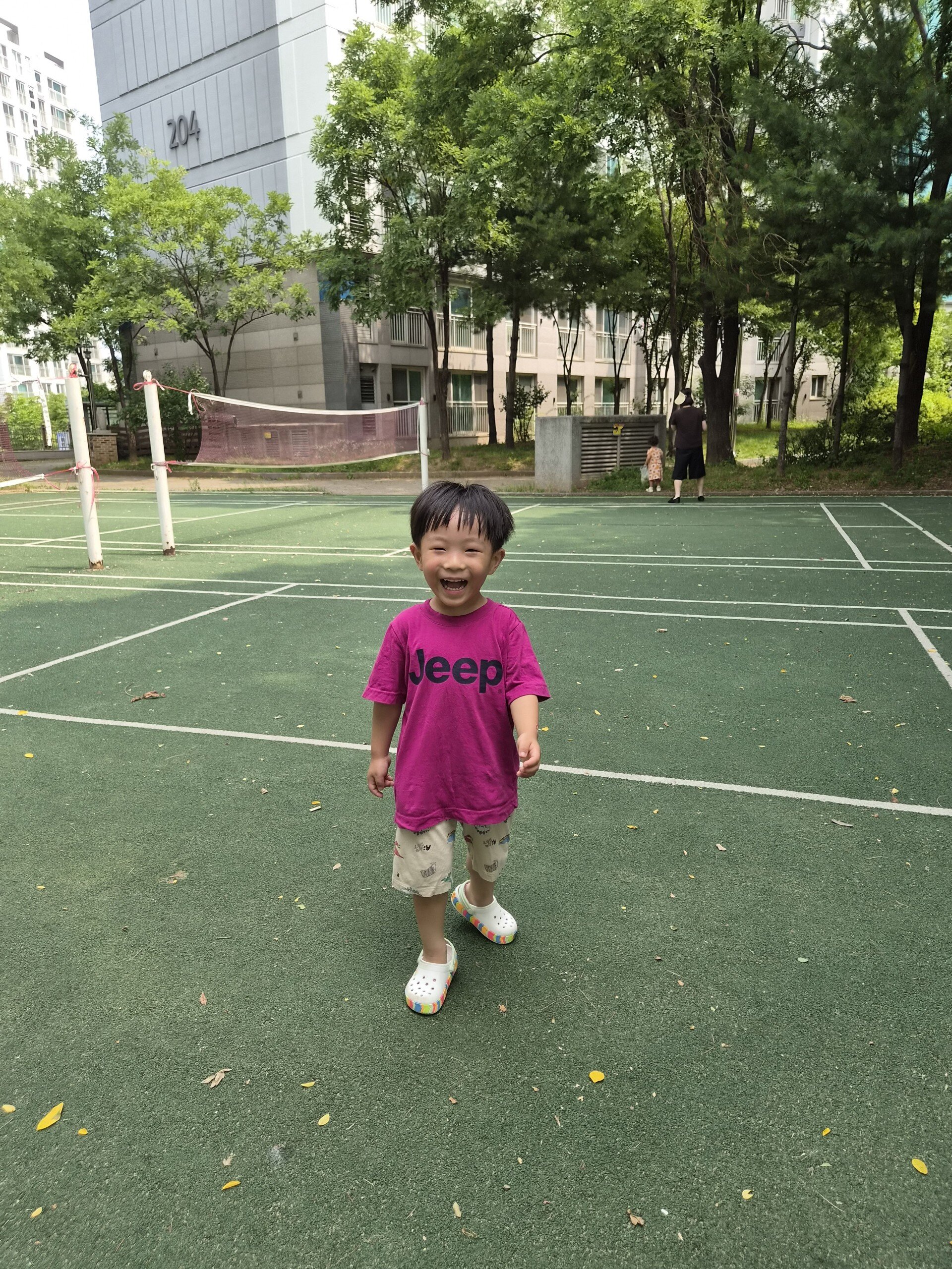 녹색 잔디 코트 위에서 환하게 웃으며 카메라를 바라보고 서 있는 지호. 뒤편에는 아파트 건물과 푸른 나무들이 보인다. 지호는 분홍색 'Jeep' 티셔츠와 짧은 바지를 입고 있다.