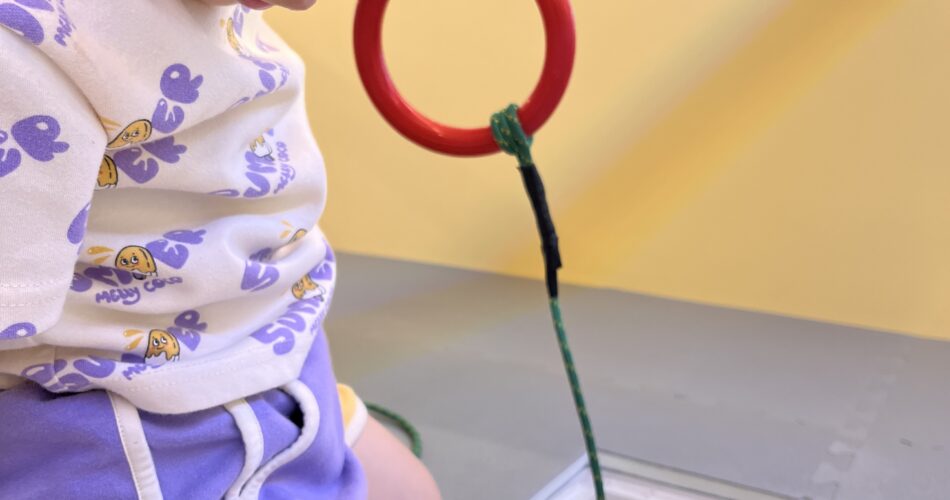하얀색 티셔츠와 보라색 반바지를 입은 지호가 무릎을 꿇고 앉아 빨간 고리 낚싯대를 이용해 투명한 상자 속 나무 고리를 낚으려 집중하고 있다.