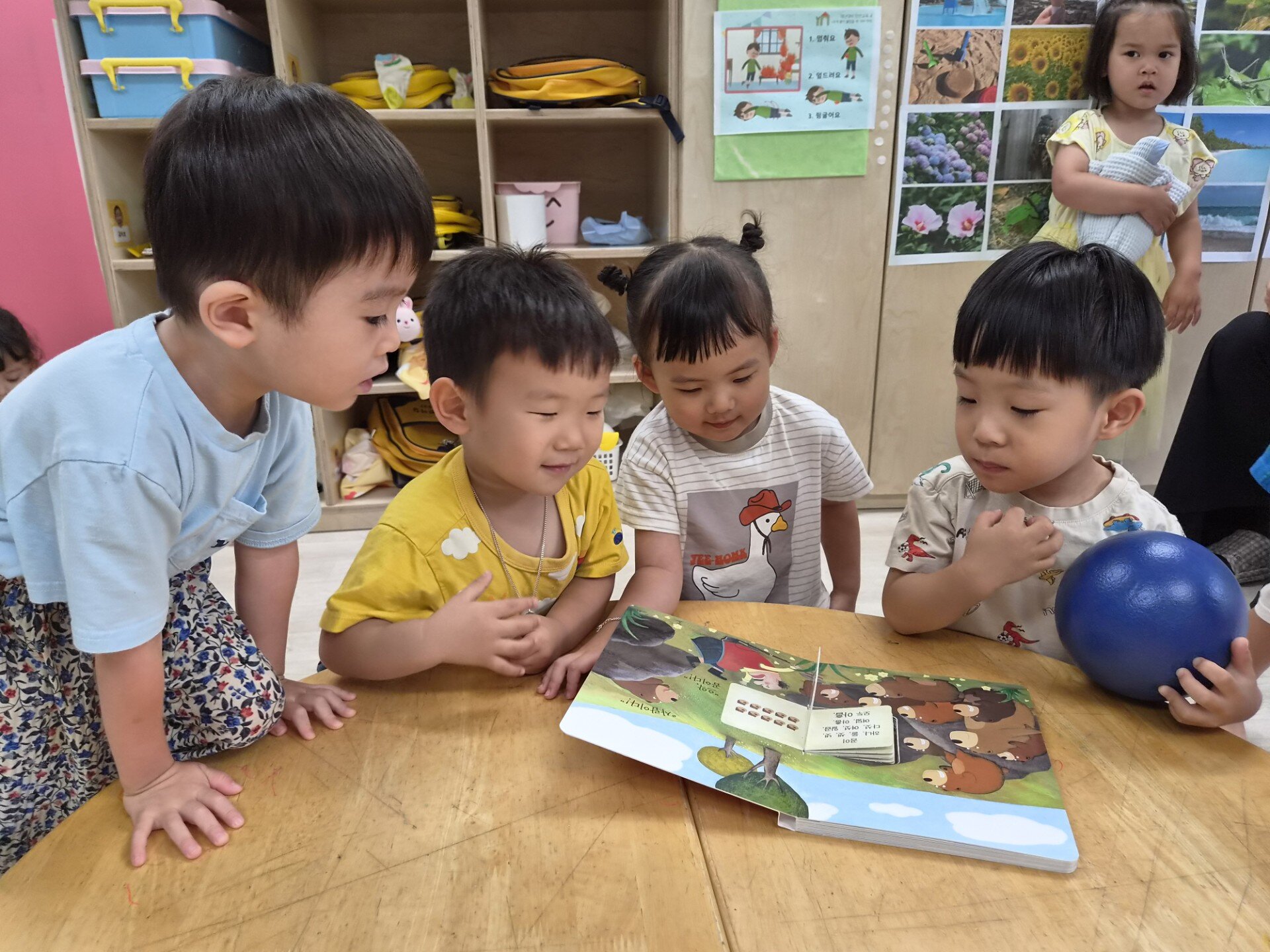 네 명의 어린이가 나무 테이블에 모여 앉아 펼쳐진 그림책을 집중해서 보고 있다. 노란색 티셔츠를 입은 지호는 오른손에 파란색 공을 들고 있으며, 뒤쪽에는 노란색 원피스를 입은 여자아이가 서 있다. 배경은 어린이집 교실 내부이다.