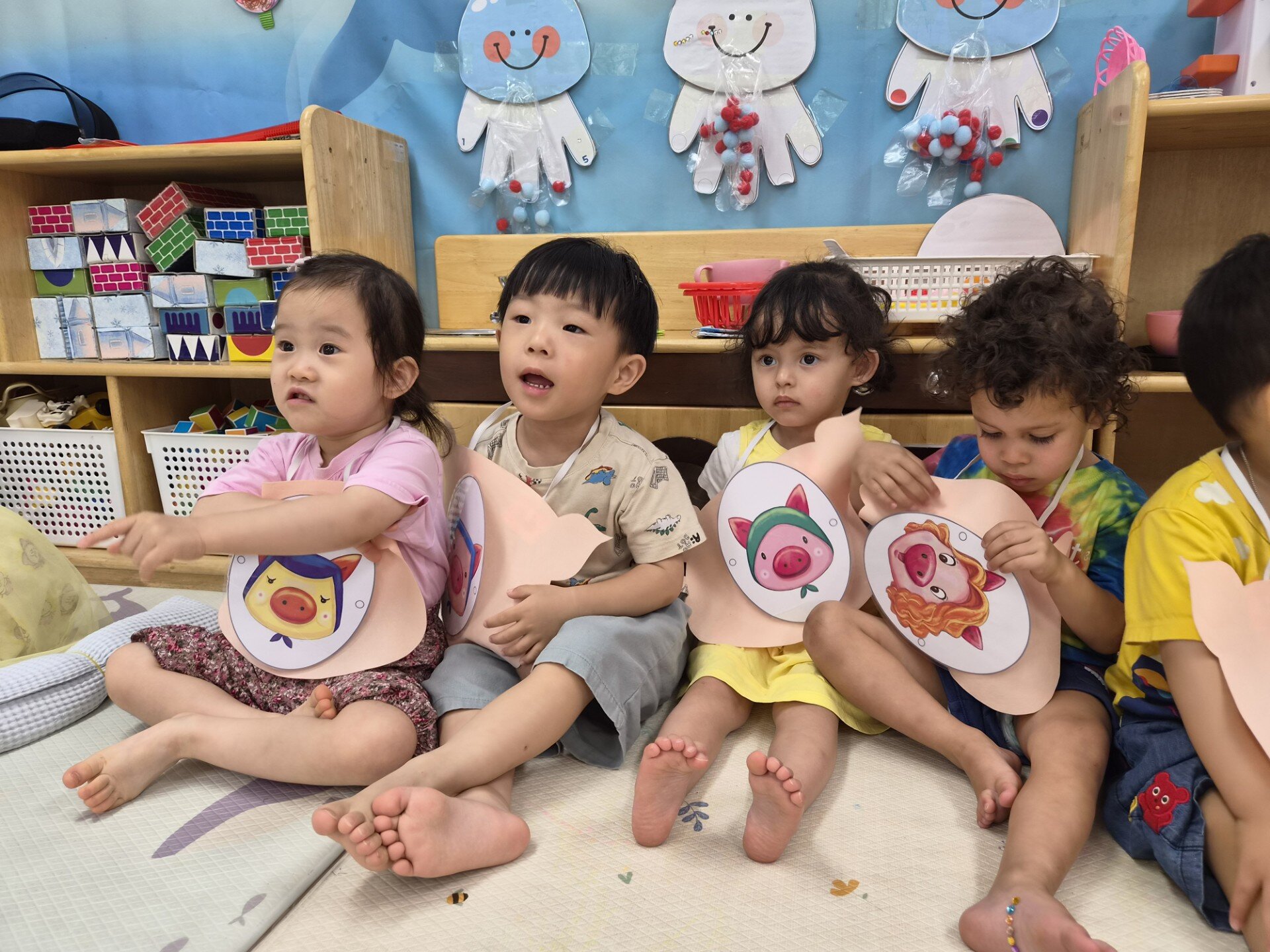 네 명의 어린이가 어린이집 교실 바닥에 앉아 돼지 캐릭터 그림이 있는 미술 활동 작품을 들고 있다. 지호는 흰색 벙거지 모자를 쓰고 중앙에 앉아있으며, 친구들과 함께 활동에 집중하는 모습이다. 배경에는 여러 교구들이 보인다.