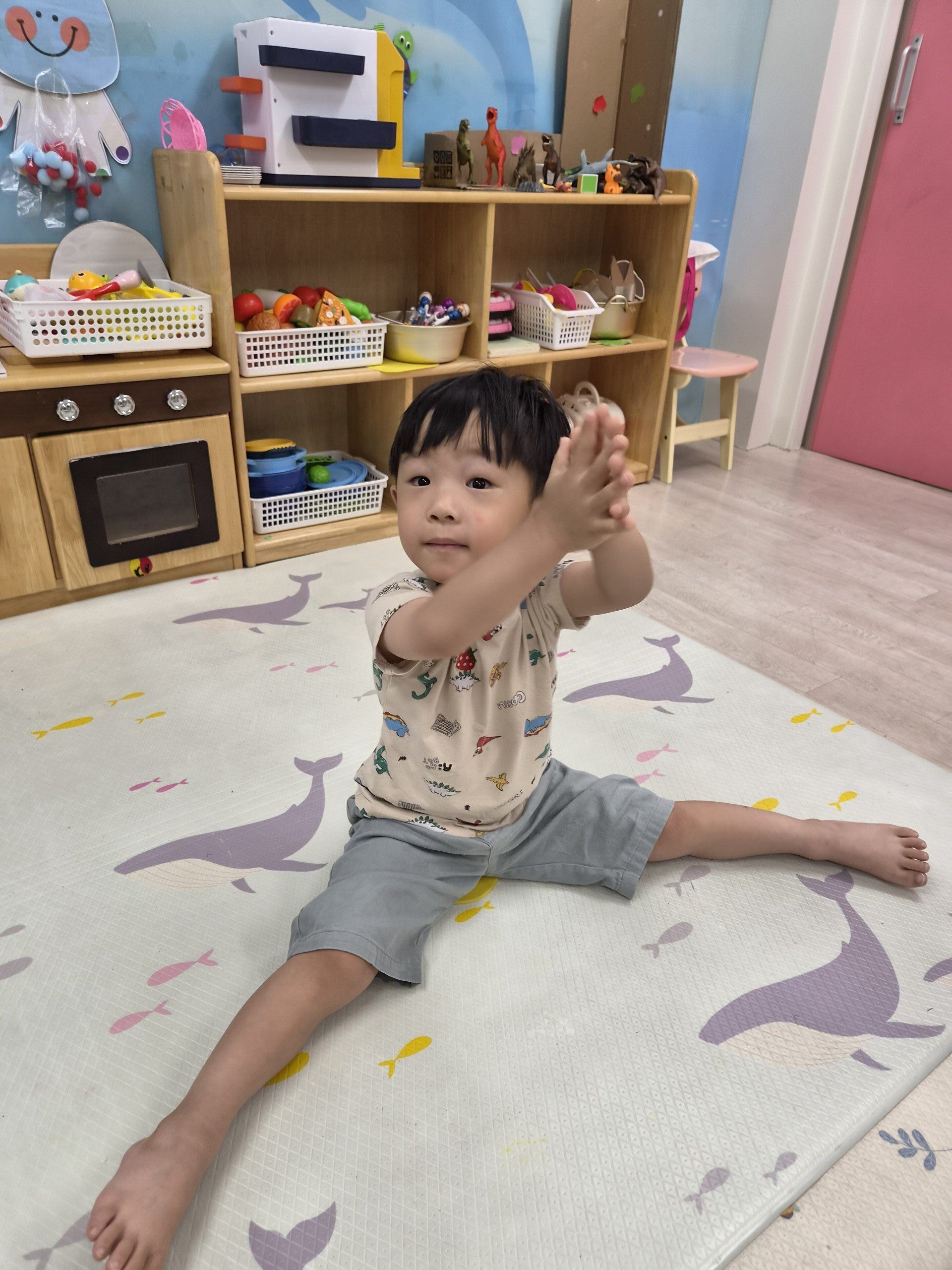 어린이집 실내 놀이방에서 지호가 고래 무늬 매트 위에서 양팔을 모으고 다리를 양옆으로 쭉 뻗어 다리 찢기를 하고 있다. 카메라를 바라보는 지호의 얼굴에 집중하는 표정과 작은 미소가 엿보인다.
