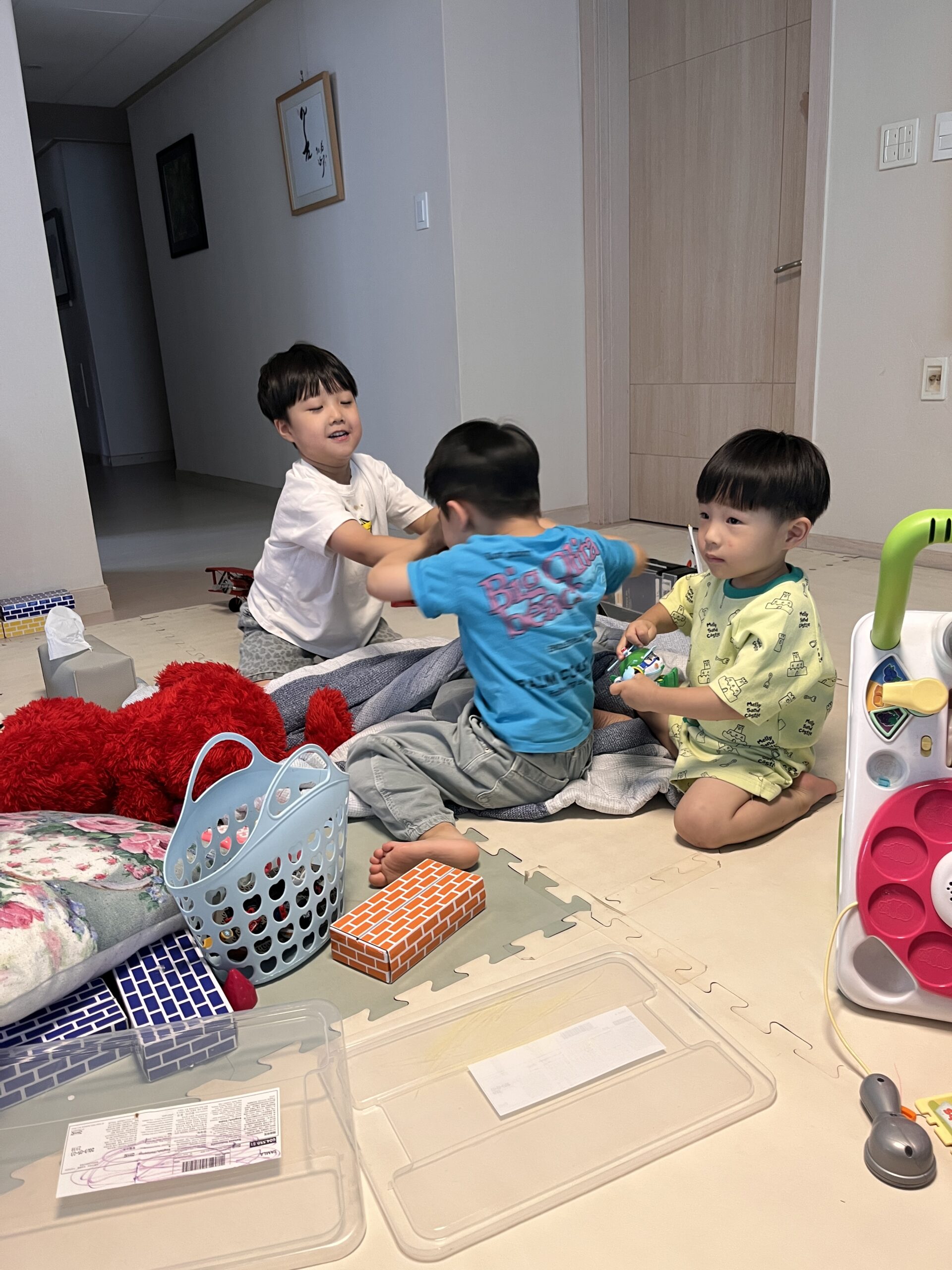 실내 거실의 놀이방 매트 위에서 세 명의 아이들이 놀고 있다. 연두색 티셔츠를 입은 지호가 바닥에 앉아 장난감을 들여다보고 있고, 다른 두 아이는 파란색 담요 위에서 몸싸움하듯 웃으며 장난치고 있다. 주변에 장난감 바구니와 블록, 쿠션 등이 널려있다.