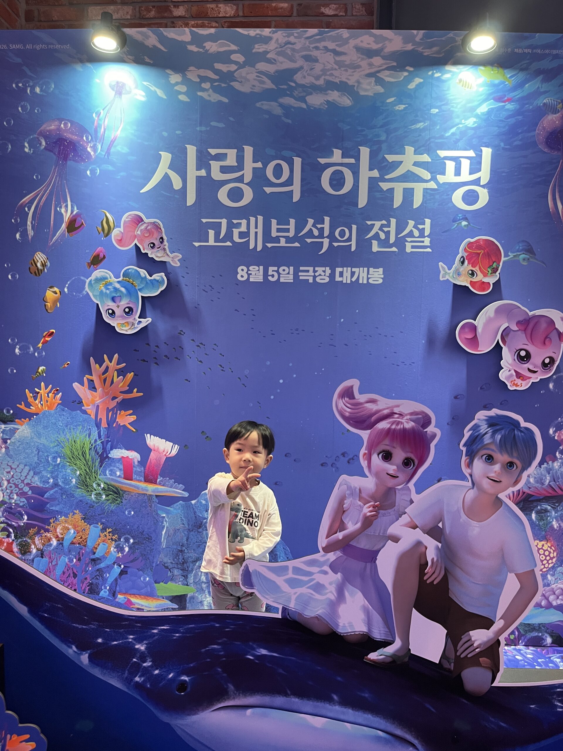 영화관으로 보이는 실내에서 지호가 '사랑의 하츄핑 고래보석의 전설' 영화 포스터 앞에서 포즈를 취하고 있다. 포스터는 바다 속 배경과 애니메이션 캐릭터들로 꾸며져 있으며, 지호는 하얀 티셔츠를 입고 손가락으로 포스터를 가리키고 있다.