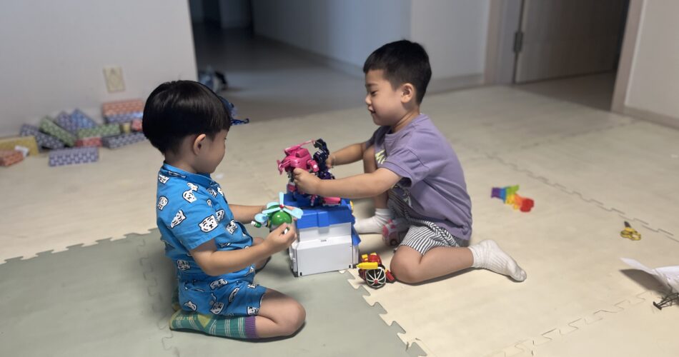 실내 매트 위에서 두 아이가 앉아 로봇 장난감과 블록을 가지고 즐겁게 놀고 있다. 왼쪽 아이는 파란색 옷을, 지호는 보라색 티셔츠를 입고 있으며, 바닥에는 다양한 색깔의 장난감들이 놓여 있다.