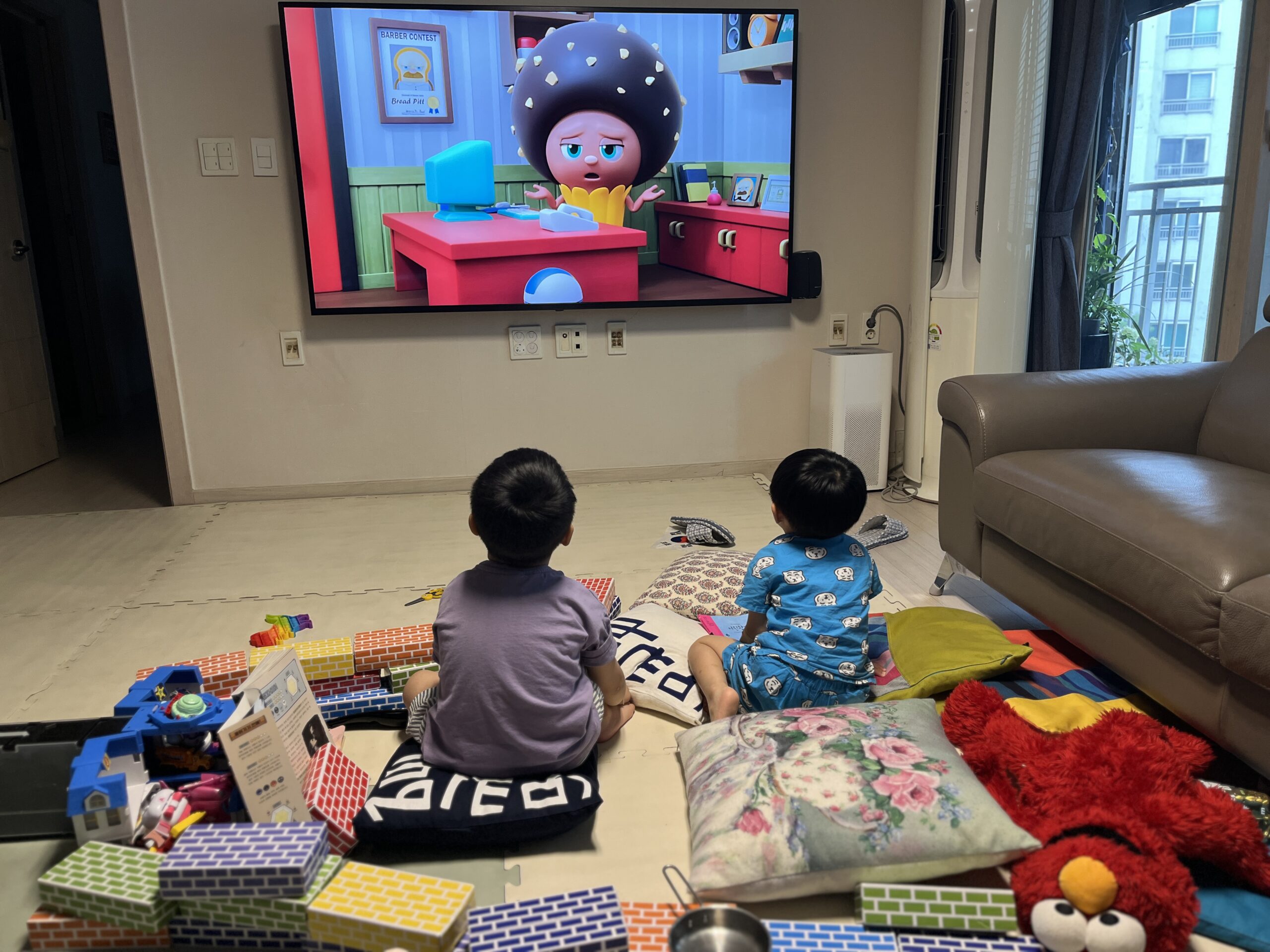 거실 바닥 매트 위에 놓인 쿠션에 파란색 상하의를 입은 지호와 보라색 티셔츠를 입은 친구가 나란히 앉아 등을 보이고 TV를 시청하고 있다. TV 화면에는 만화 캐릭터가 보이며, 주변에는 여전히 블록과 장난감들이 놓여있다.
