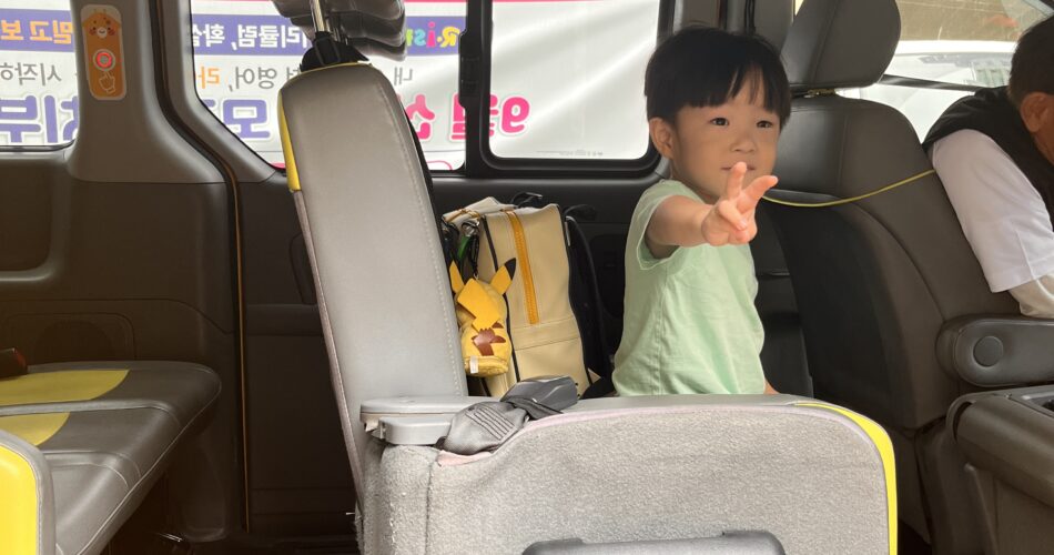 밝은 녹색 티셔츠를 입은 지호가 노란색 좌석이 있는 차량 안에서 카메라를 향해 손가락을 뻗고 있다. 뒤에는 노란색 배낭이 놓여 있고, 창밖에는 건물이 비친다.