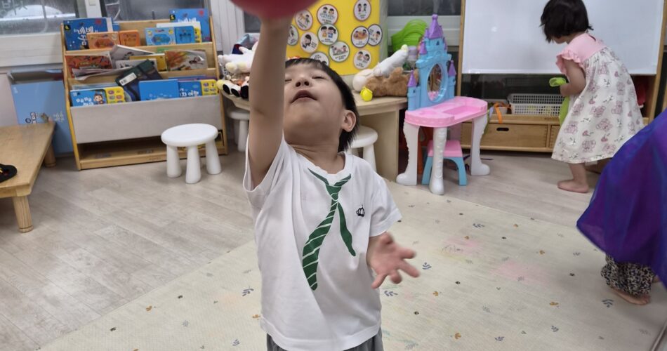 밝은 어린이집 교실에서 지호가 맨발로 서서 빨간색 공을 향해 오른팔을 높이 뻗어 잡으려 하고 있다. 티셔츠에는 녹색 넥타이 무늬가 그려져 있고, 주변에는 책장과 장난감들이 보인다.