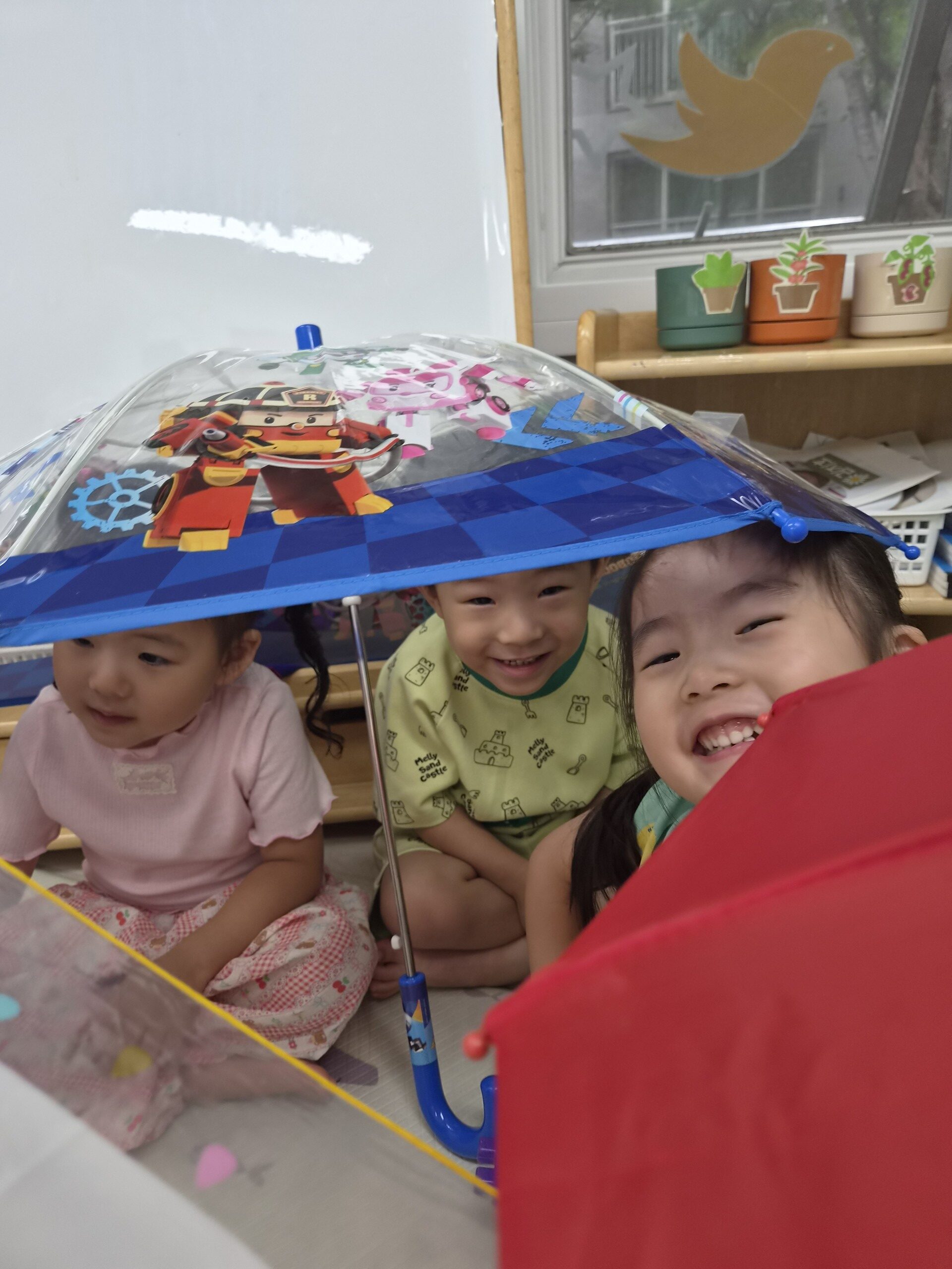 실내에서 세 명의 아이들이 투명한 어린이 우산 아래에 옹기종기 앉아 밝게 웃고 있다. 정면에 있는 아이는 크게 웃으며 카메라를 보고 있고, 뒤편에는 캐릭터 그림이 그려진 우산이 보인다.