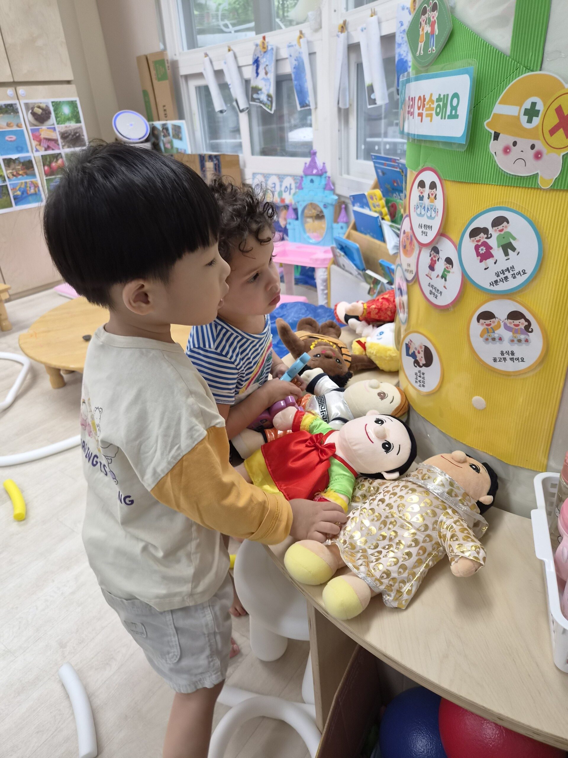 지호와 다른 남자아이가 어린이집 선반에 놓인 여러 인형과 벽에 붙은 교육용 약속 그림판을 흥미롭게 구경하고 있습니다. 지호는 한복 인형 옆에 서서 위를 올려다보고 있습니다.