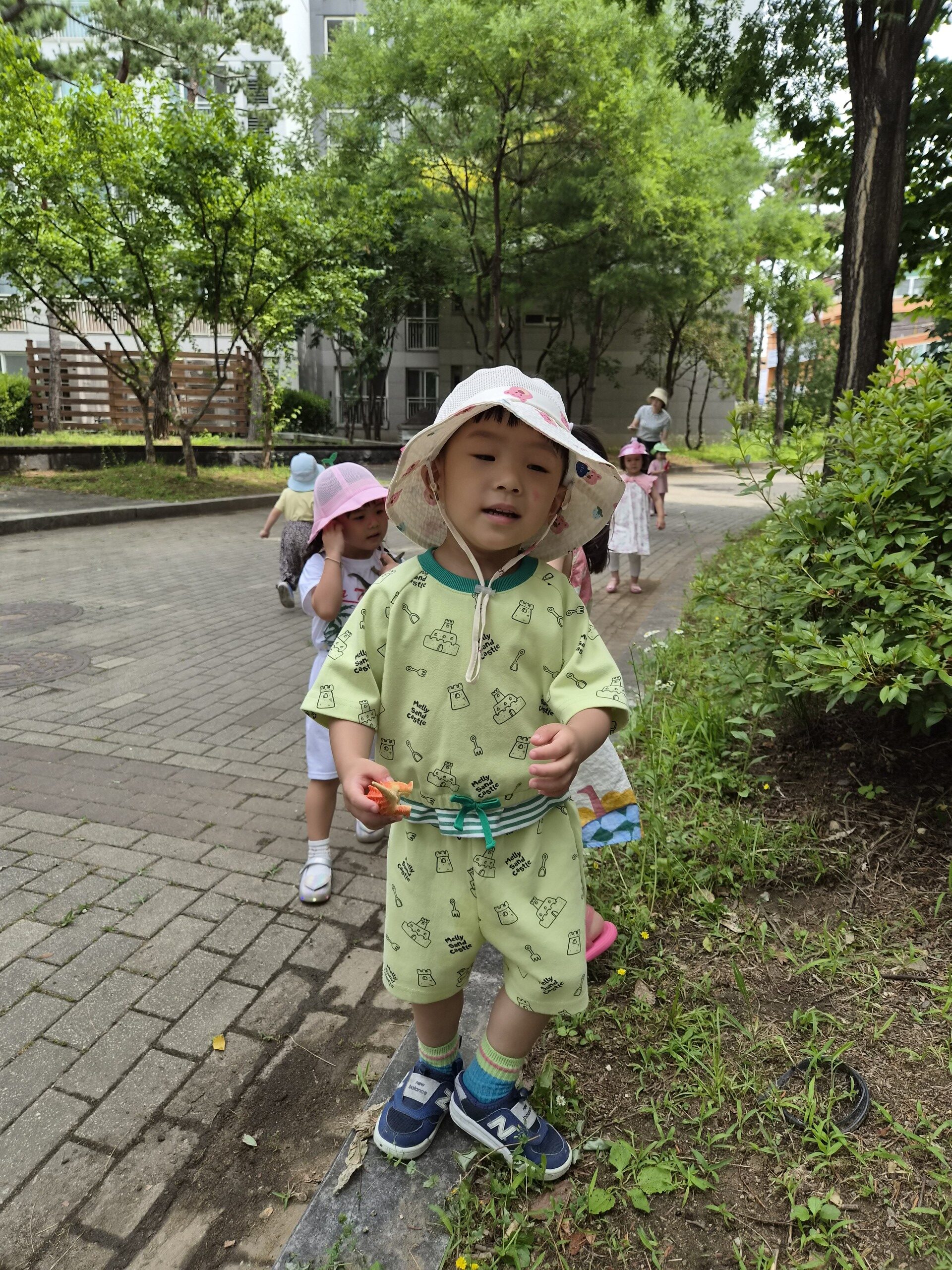 나무와 풀이 우거진 길을 어린이집 친구들과 함께 걷고 있는 지호. 연두색 반팔 상하의와 흰색 버킷햇을 쓰고 작은 주황색 장난감을 쥐고 카메라를 응시하고 있다. 뒤편에는 다른 아이들이 줄지어 걸어오고 있다.