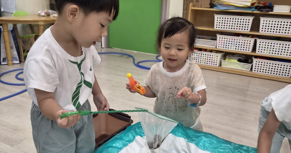 어린이집 실내에서 지호와 여자아이가 낮은 초록색 물놀이 풀에 앉아 물고기 장난감을 잡으려 그물망과 노란색 장난감을 들고 집중하고 있다. 주변에 다양한 색깔의 물놀이 장난감들이 보인다.
