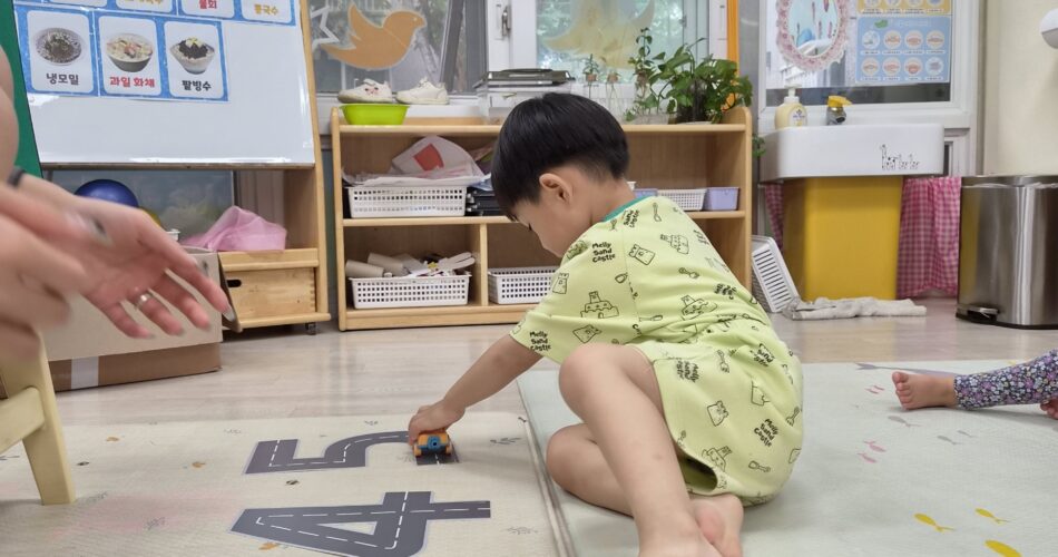 밝은 실내 어린이집 교실 바닥에 앉아 도로 매트 위에서 작은 파란색 장난감 자동차를 밀며 놀고 있는 지호의 모습. 왼쪽에 어른의 손 일부가 보인다. 지호는 연두색 상하복을 입고 맨발이다.
