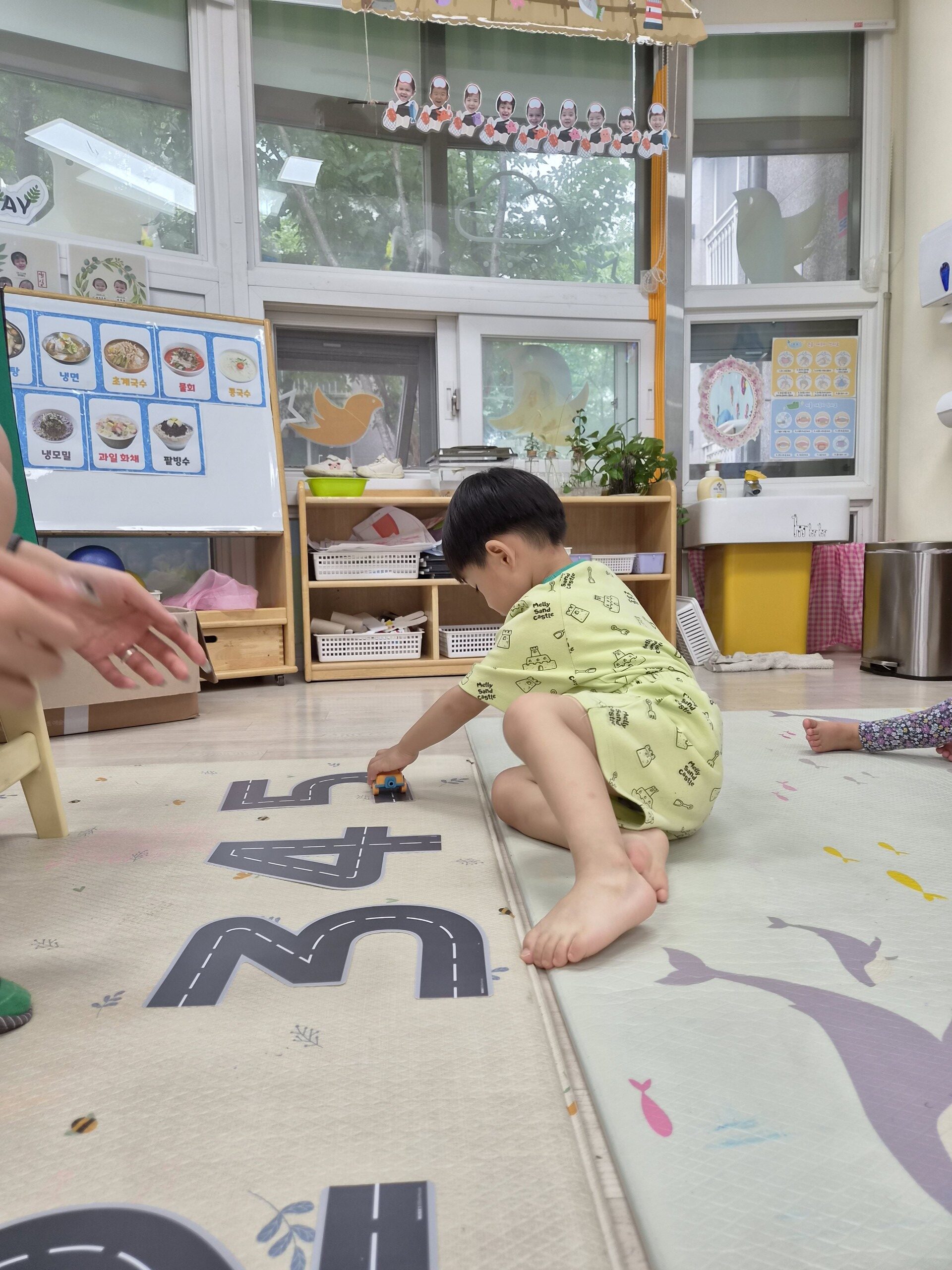 밝은 실내 어린이집 교실 바닥에 앉아 도로 매트 위에서 작은 파란색 장난감 자동차를 밀며 놀고 있는 지호의 모습. 왼쪽에 어른의 손 일부가 보인다. 지호는 연두색 상하복을 입고 맨발이다.