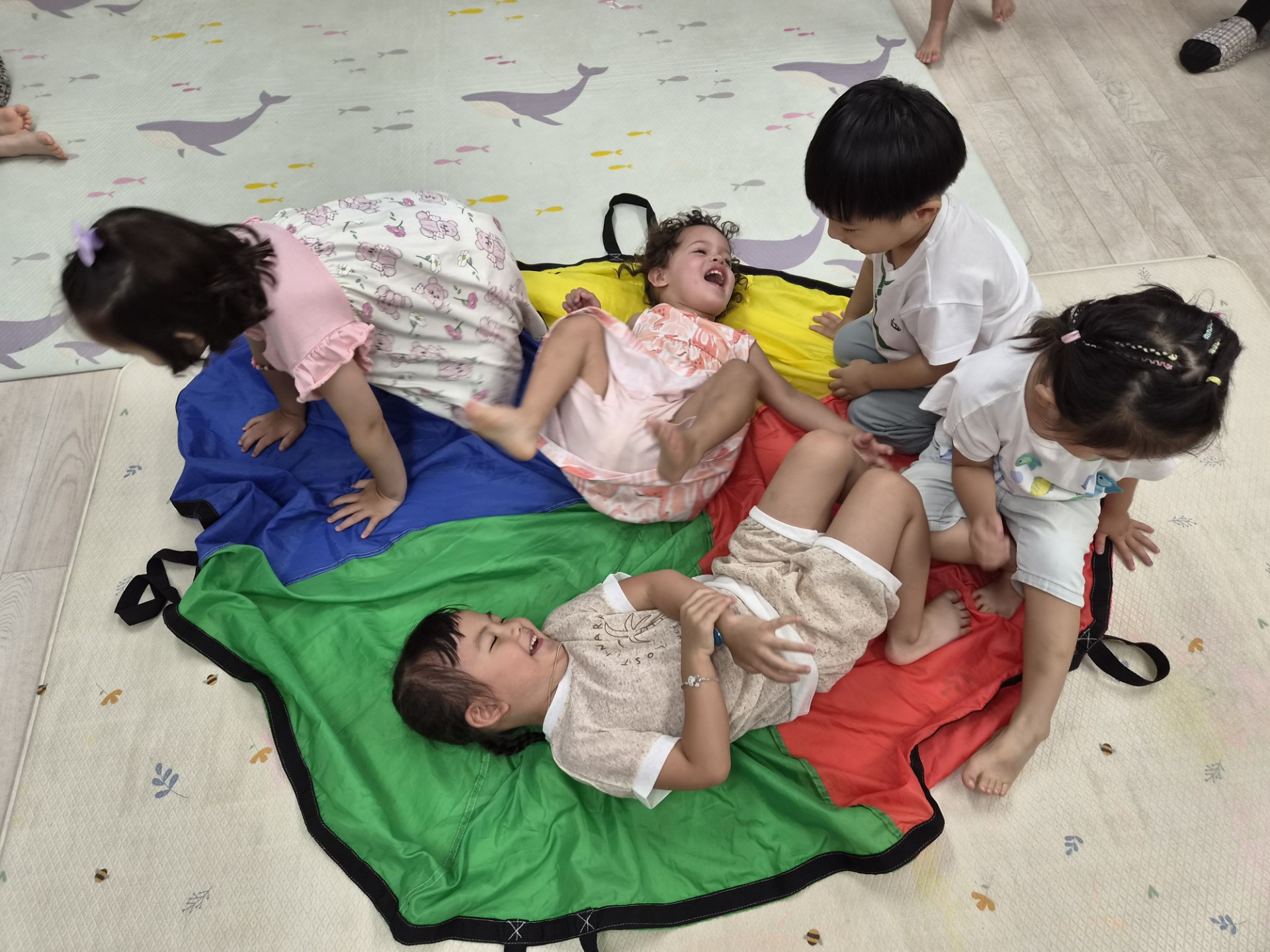 여러 명의 아이들이 형형색색의 낙하산 천 위에서 활기차게 놀고 있다. 두 아이는 천 위에 누워 환하게 웃고 있고, 다른 아이들은 주변에 앉거나 엎드려 즐거운 시간을 보내고 있다.