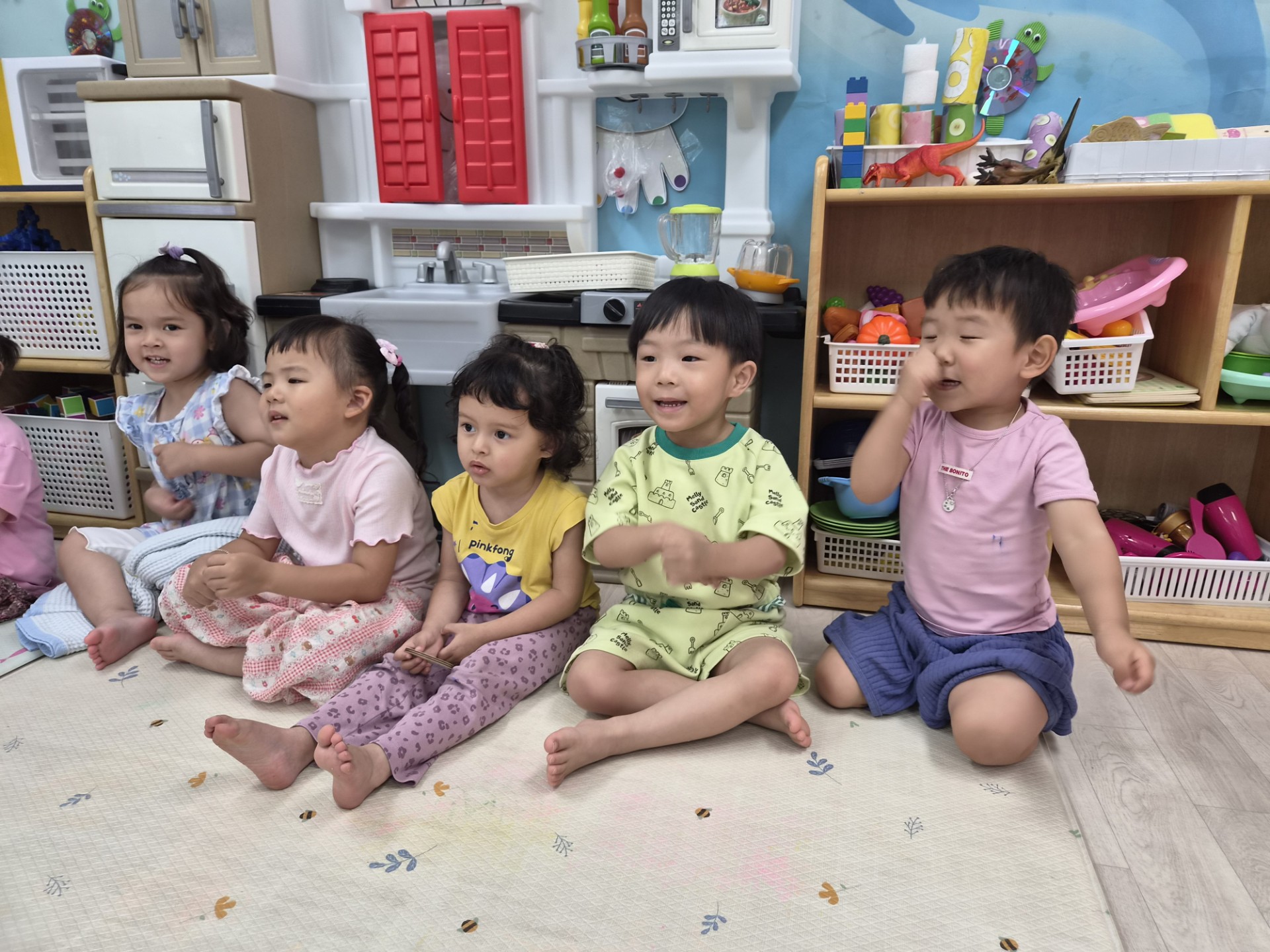 다섯 명의 아이들이 실내 바닥에 앉아 정면을 바라보며 활동에 참여하고 있다. 한 아이는 코를 만지고 있고, 다른 아이들은 집중한 표정이다. 뒤쪽으로는 주방 놀이 세트가 보인다.