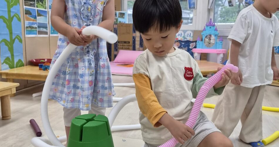 어린이집 실내 놀이 공간에서 지호와 여자아이가 초록색 원뿔 모양 블록에 흰색과 분홍색 구불구불한 튜브를 연결하고 있습니다. 지호는 황토색과 연두색 소매가 배색된 티셔츠를 입고 있습니다.
