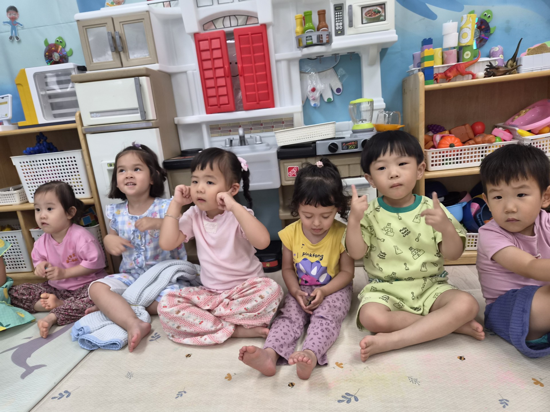 다섯 명의 아이들이 실내 바닥에 옹기종기 모여 앉아 정면을 바라보고 있다. 아이들은 진지한 표정으로 무언가에 집중하고 있으며, 뒤편에는 아기자기한 교구장과 주방 놀이 세트가 보인다.