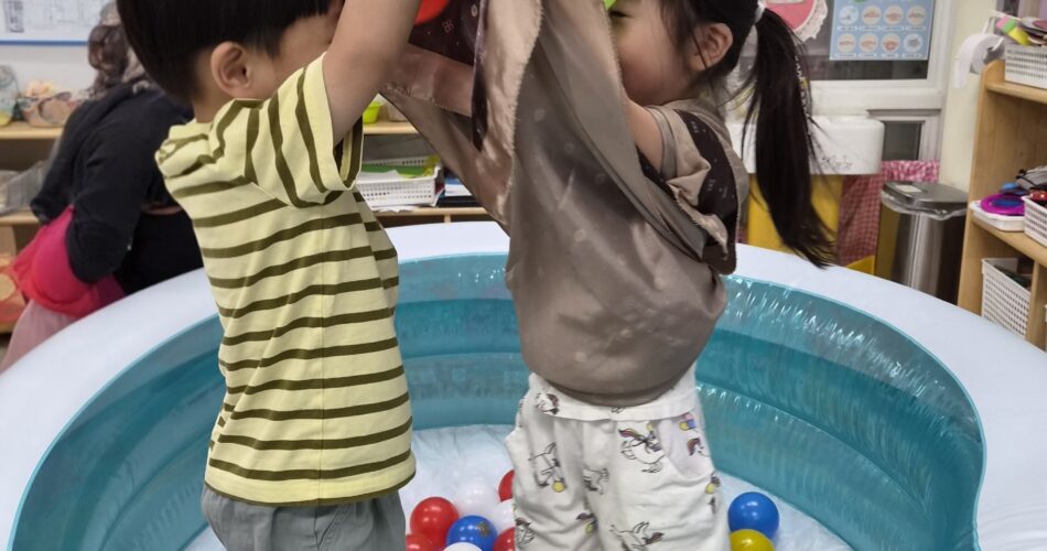 노란색 줄무늬 티셔츠를 입은 지호와 다른 여자아이가 파란색 공기 주입식 볼풀 안에서 장난감을 함께 잡고 놀고 있다. 주변에는 형형색색의 공들이 가득하다.