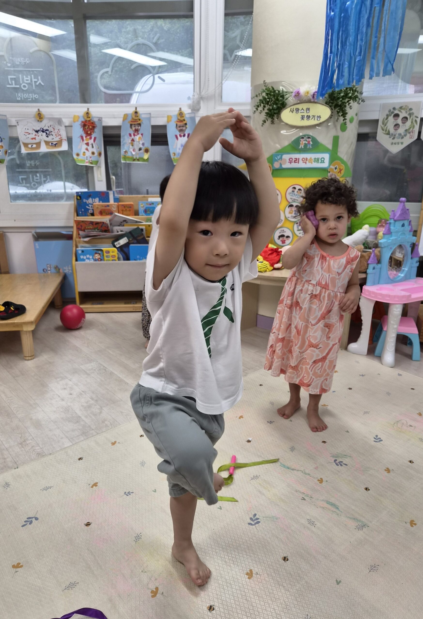 어린이집 교실에서 지호가 맨발로 한쪽 다리를 들고 두 팔을 머리 위로 올려 균형을 잡고 있다. 카메라를 향해 장난기 가득한 미소를 짓고 있으며, 뒤편에는 다른 친구가 서 있다.