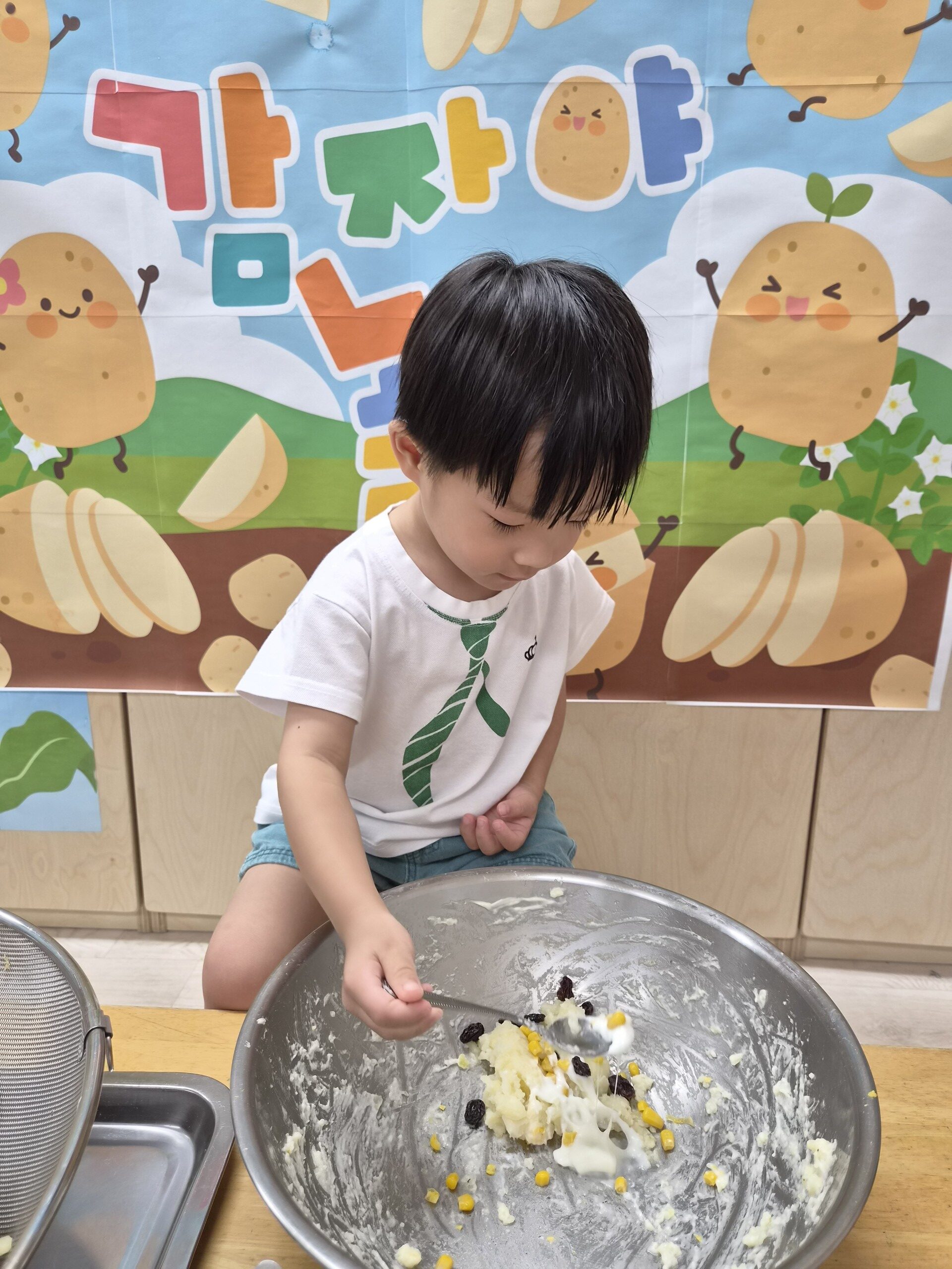 하얀색 티셔츠에 녹색 넥타이 무늬가 프린트된 옷을 입은 지호가 주방 조리대 앞에서 무릎을 꿇고 앉아 스테인리스 볼에 담긴 감자 샐러드 재료(으깬 감자, 옥수수, 건포도 등)를 숟가락으로 섞고 있는 모습. 뒤편에는 '감자야'라고 적힌 귀여운 배경 현수막이 보인다.
