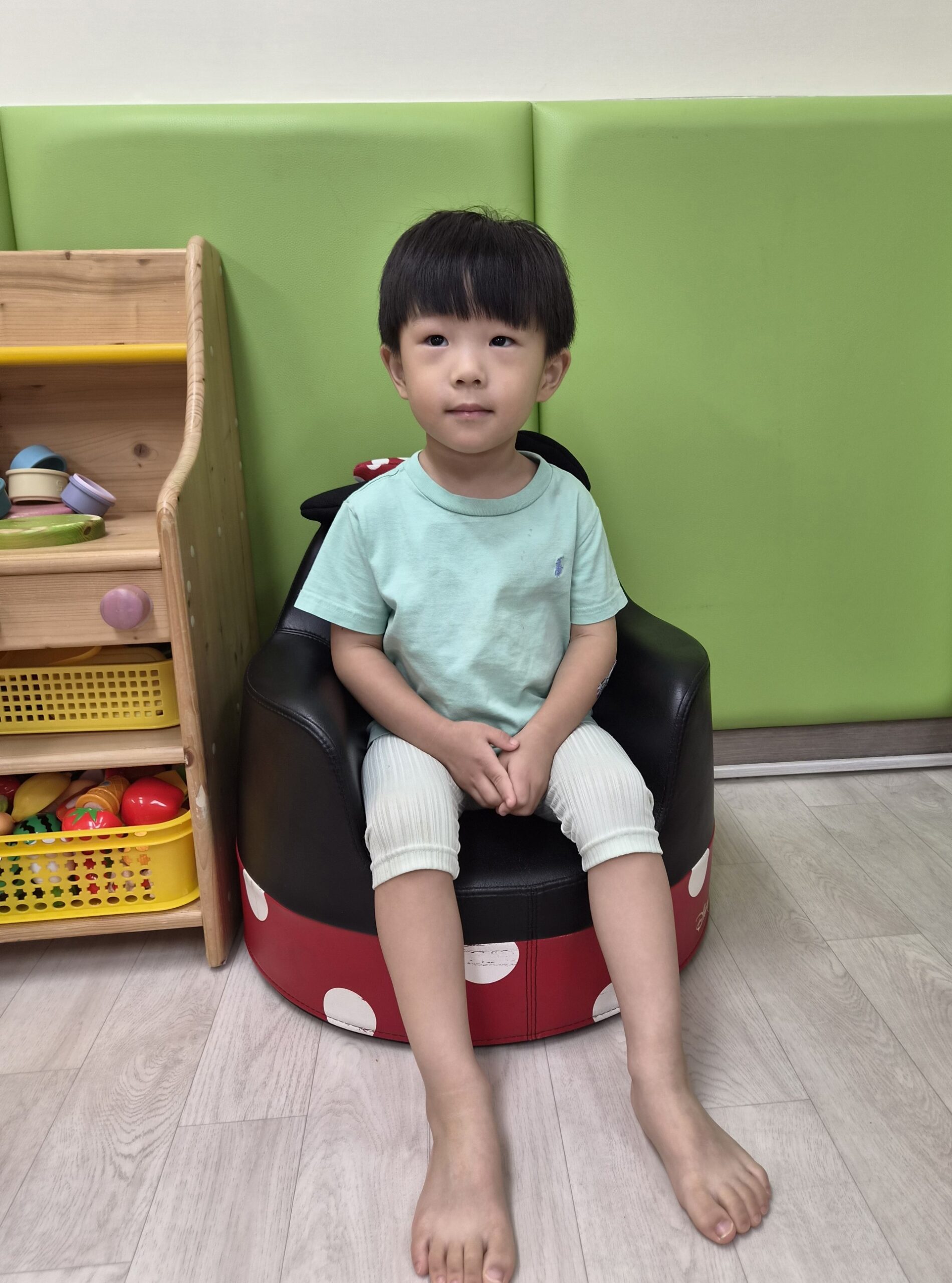 지호가 민트색 티셔츠와 밝은 색 바지를 입고 귀여운 캐릭터 모양의 검은색 어린이 의자에 맨발로 앉아 카메라를 응시하고 있다. 배경에는 초록색과 흰색 벽, 장난감 선반이 보인다.