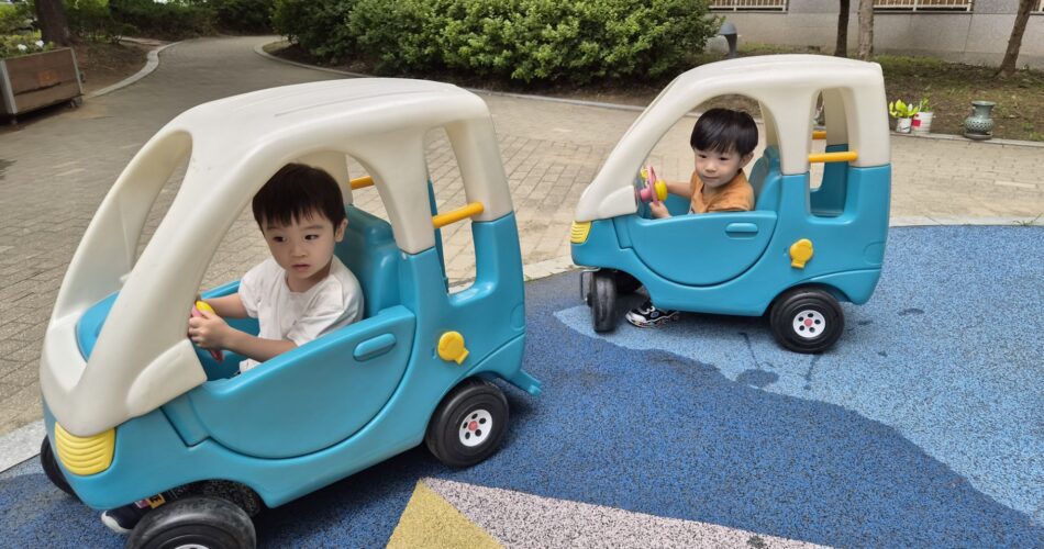 야외 놀이터에서 파란색과 흰색의 유아용 장난감 자동차에 앉아 운전대를 잡고 있는 지호와 다른 아이의 모습. 주변에는 푸른 나무와 아파트 건물이 보인다.