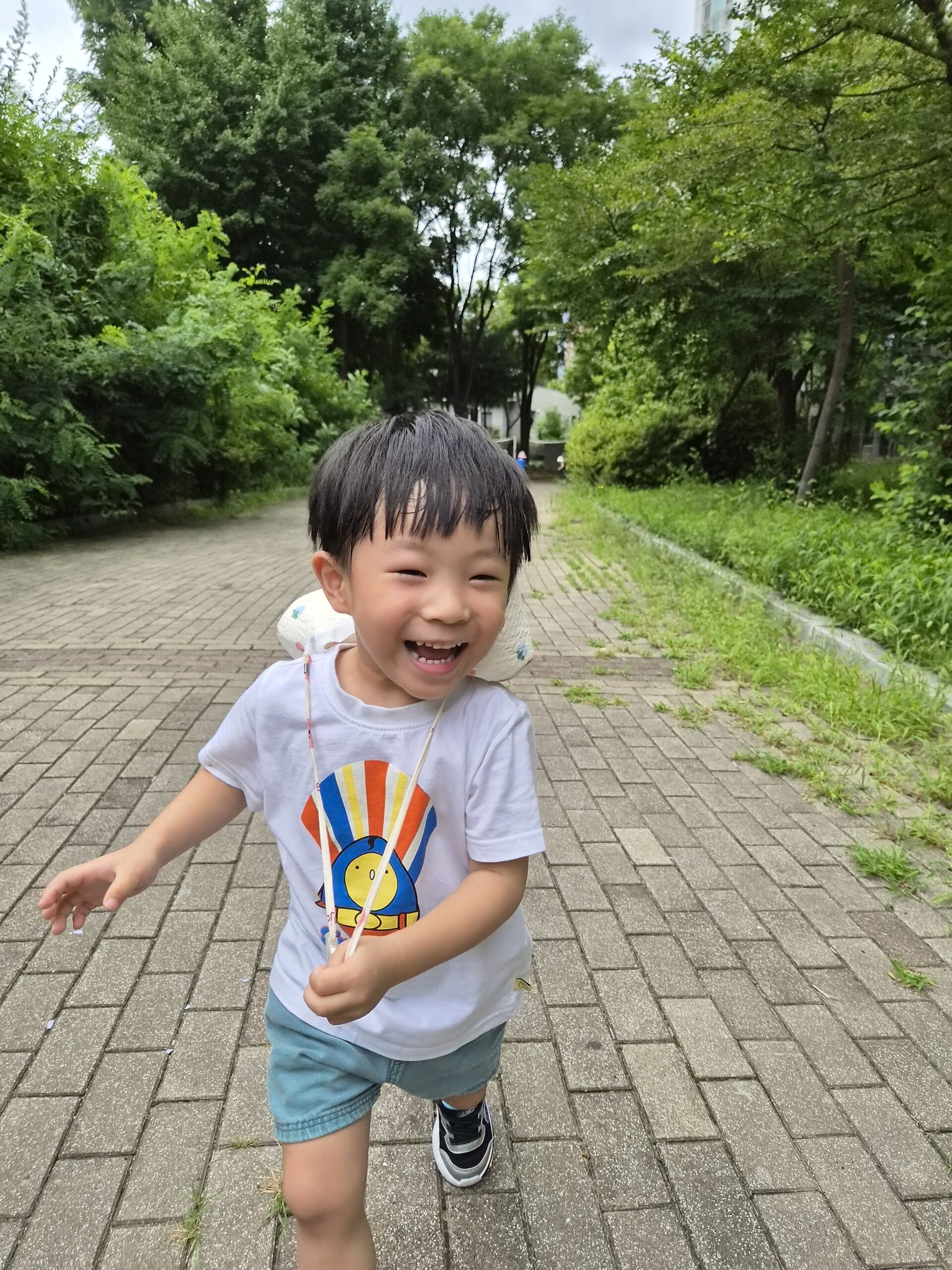 야외 포장된 길 위에서 해맑게 웃으며 뛰어오는 지호. 왼쪽 팔을 살짝 들고 앞을 향해 나아가고 있으며, 뒤편으로는 푸른 나무들이 이어져 있다. 여전히 흰색 캐릭터 티셔츠와 파란색 반바지를 입고 있다.