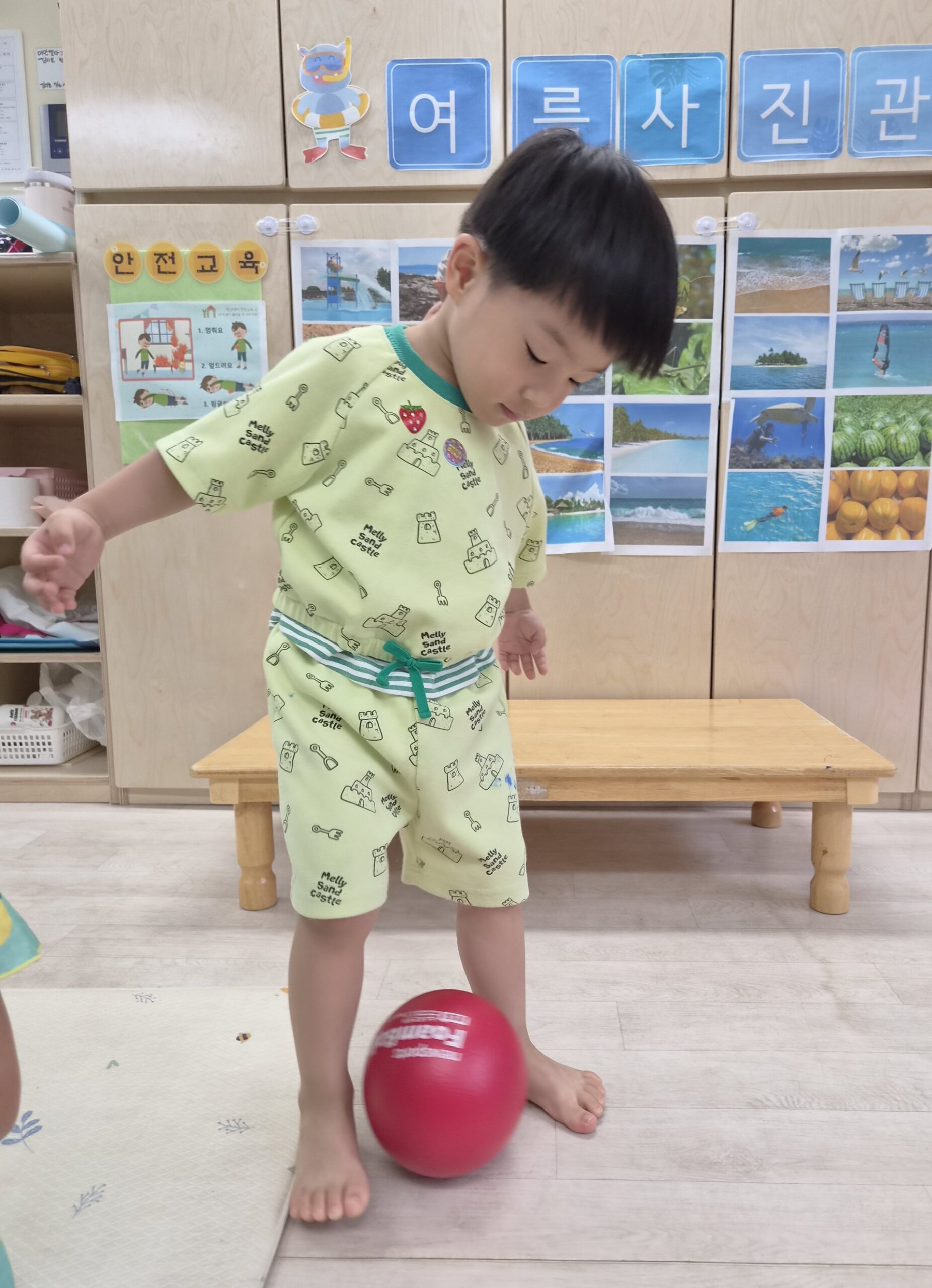연두색 상하의를 입은 맨발의 지호가 빨간색 공을 발로 살짝 건드리며 아래를 내려다보고 있다. 뒤편에는 '여름 사진관'이라는 글씨와 함께 바다 풍경 사진들이 붙어 있다.