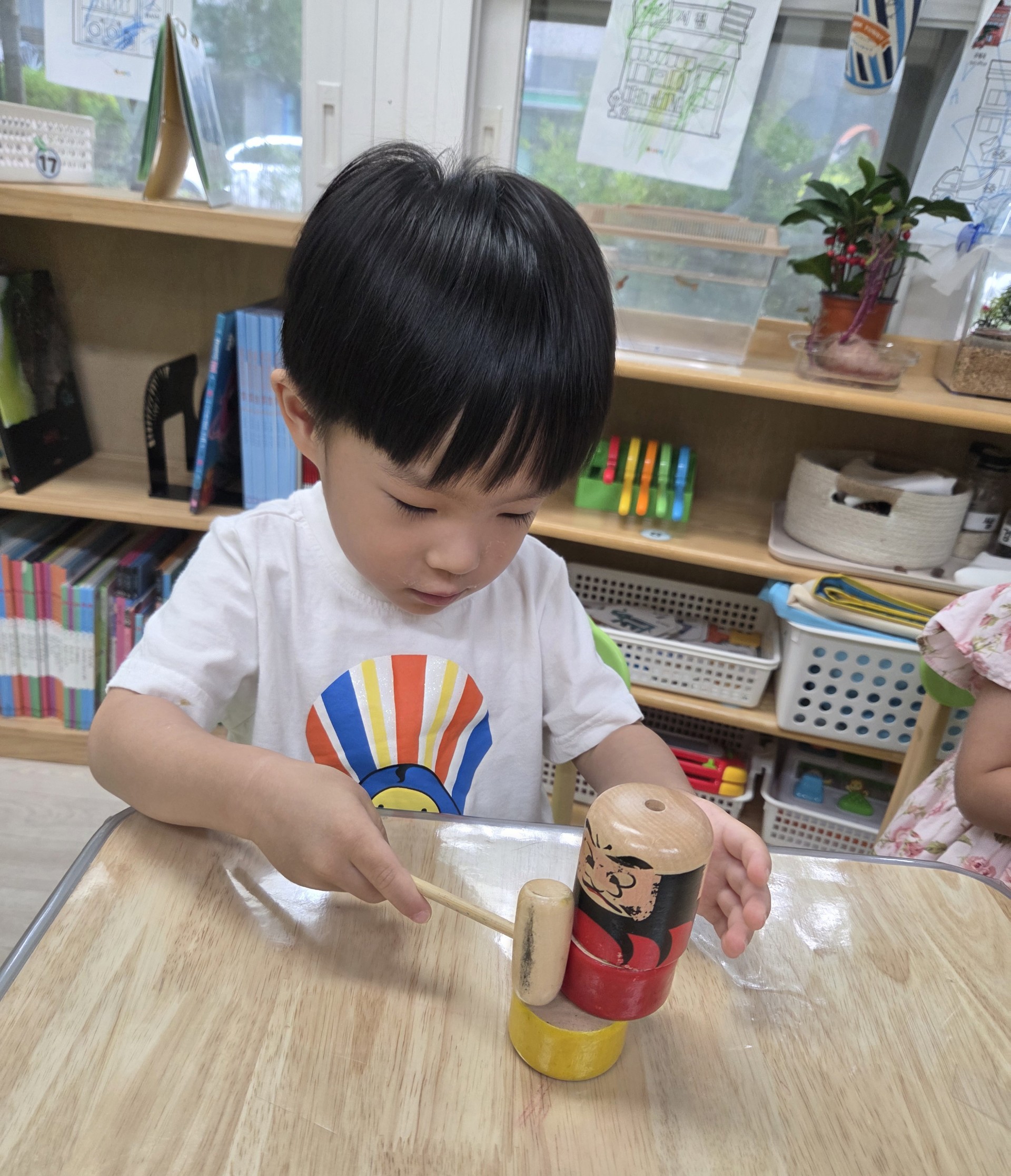 지호가 테이블에 앉아 작은 나무 망치로 사람 모양의 나무 장난감을 두드리고 있다. 배경에는 다양한 교구들이 정리된 선반이 보인다.