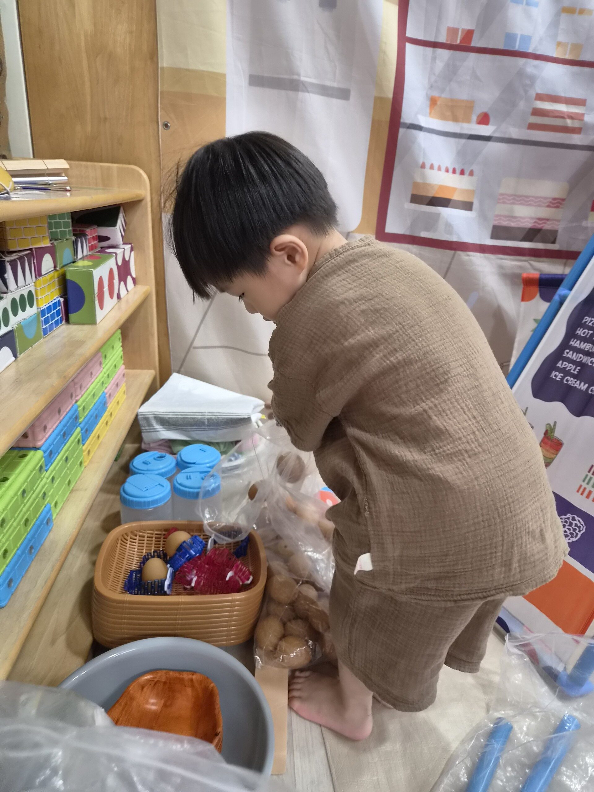 갈색 옷을 입은 지호가 선반 앞에 쪼그리고 앉아 감자처럼 보이는 재료들이 담긴 봉투와 바구니를 유심히 살펴보고 있다. 선반 위에는 블록과 물병 등이 놓여 있다.