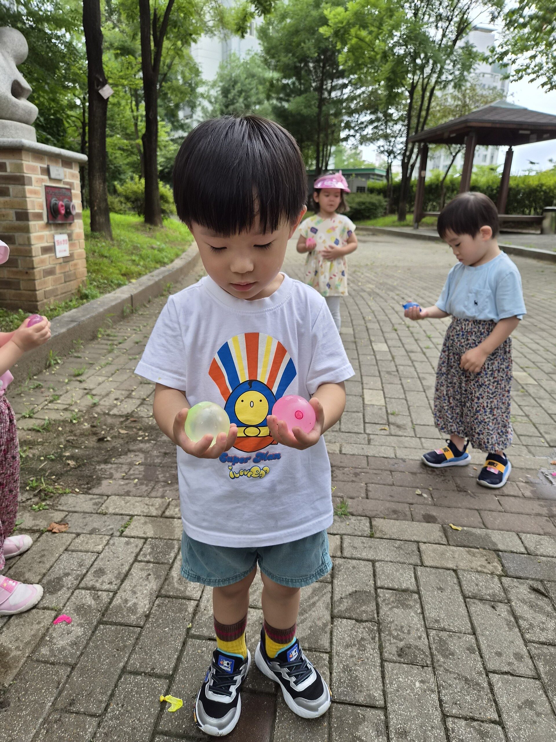 야외 보도블록 위에서 지호가 하얀색 티셔츠와 파란색 반바지를 입고 서서 양손으로 핑크색과 연두색 물풍선 두 개를 신중하게 들여다보고 있다. 주변에는 다른 아이들도 함께 있다.