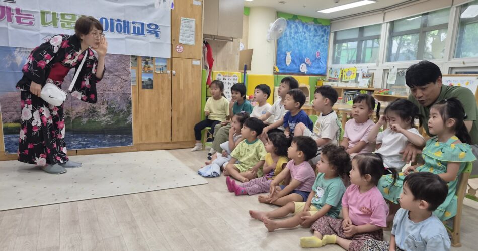 화려한 기모노를 입은 선생님이 아이들 앞에서 손짓하며 이야기하고 있고, 여러 명의 아이들이 바닥에 앉아 집중해서 선생님을 바라보고 있다. 배경에는 '다문화 이해교육'이라는 글씨가 적힌 현수막이 보인다.