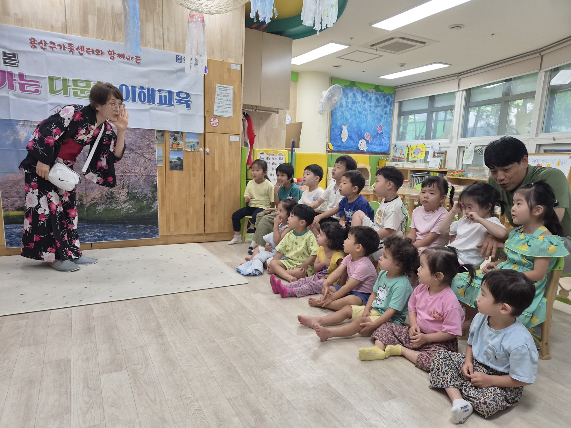 화려한 기모노를 입은 선생님이 아이들 앞에서 손짓하며 이야기하고 있고, 여러 명의 아이들이 바닥에 앉아 집중해서 선생님을 바라보고 있다. 배경에는 '다문화 이해교육'이라는 글씨가 적힌 현수막이 보인다.