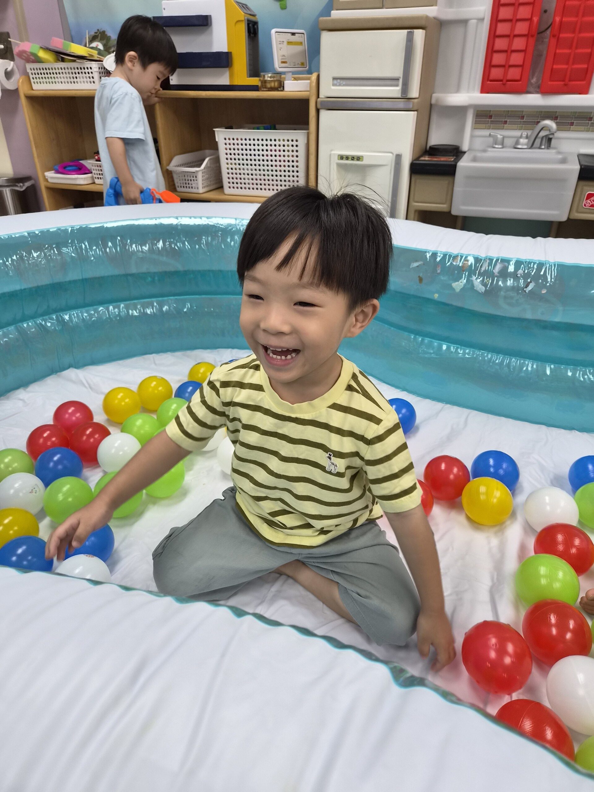 파란색 볼풀장 안에 앉아있는 지호가 환하게 웃으며 카메라를 바라보고 있다. 그의 주변에는 빨강, 노랑, 초록, 파랑 등 다양한 색깔의 공들이 가득하다.