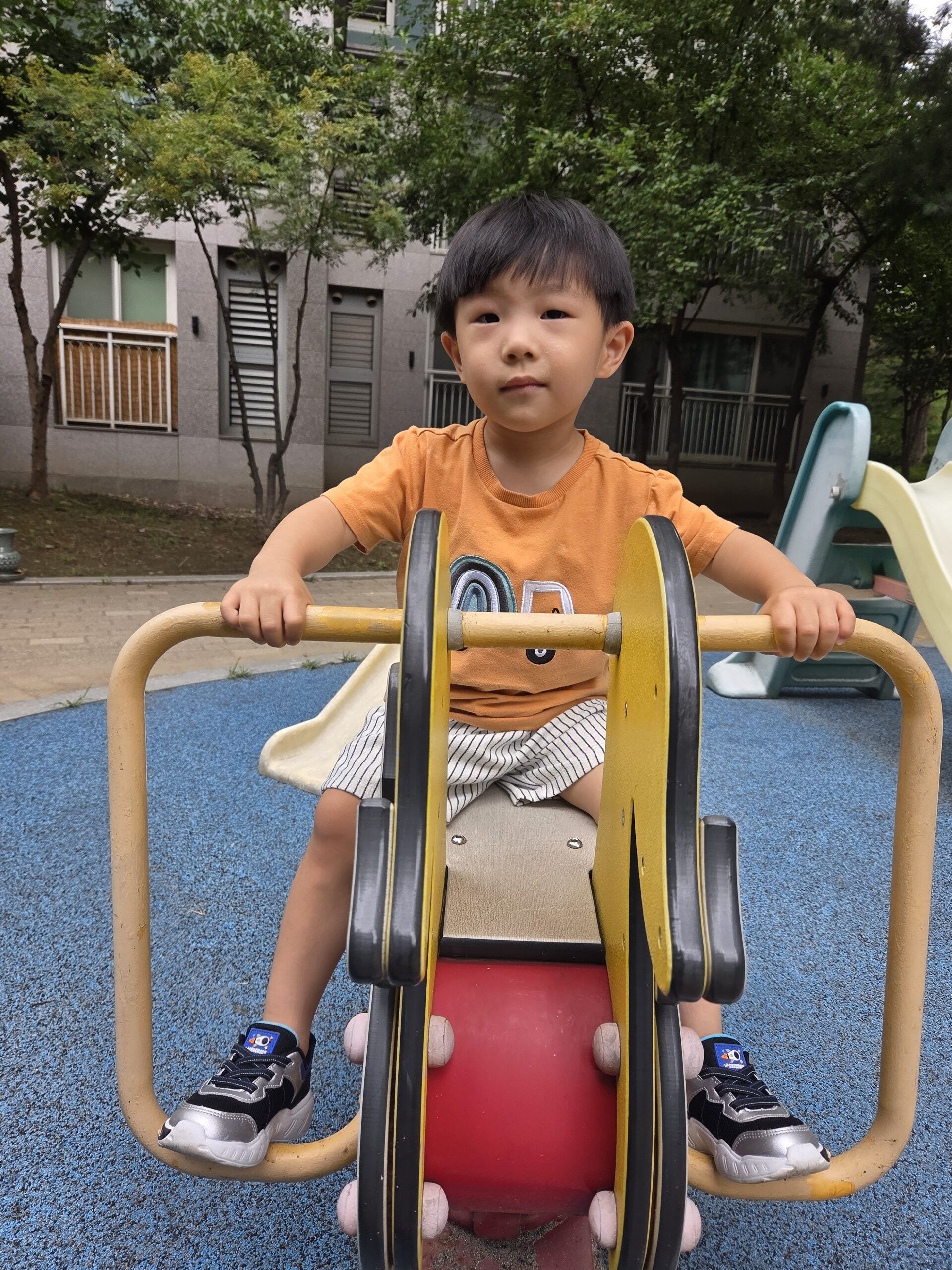 노란색 오리 스프링 흔들말에 앉아 정면을 응시하고 있는 지호의 모습. 주황색 티셔츠를 입고 양손으로 손잡이를 꽉 잡고 있다. 뒤로는 아파트 건물과 나무, 그리고 미끄럼틀이 보인다.