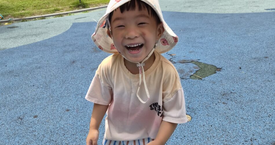 파란색 고무 바닥 놀이터에서 흰색 버킷햇과 줄무늬 바지를 입은 지호가 카메라를 향해 입을 크게 벌리고 환하게 웃고 있다. 뒤쪽으로는 푸른 잔디밭과 아파트 건물이 보인다.