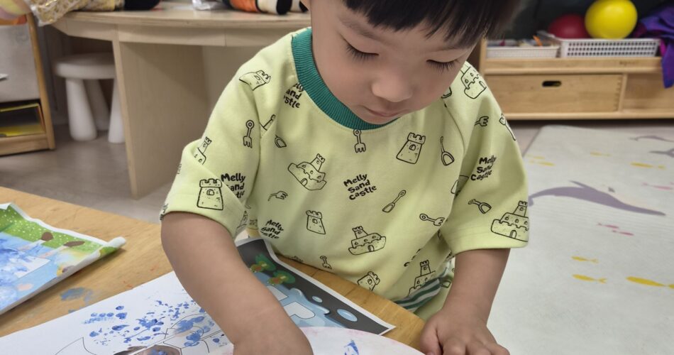 연두색 티셔츠를 입은 어린 남자아이 지호가 나무 탁자에 앉아 파란색 물감을 듬뿍 담은 종이 접시 위에서 오른손 검지 손가락으로 물감을 휘젓고 있는 모습. 접시 옆에는 밑그림이 그려진 종이가 놓여 있다.