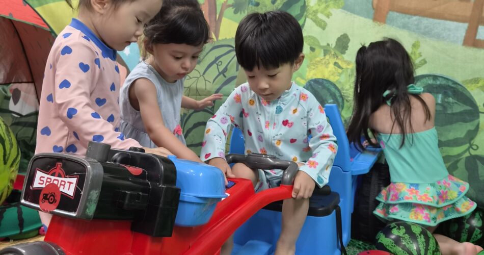 지호가 수박 테마로 꾸며진 실내 공간에서 다른 아이들과 함께 빨간색과 파란색 장난감 자동차를 타고 노는 모습. 지호는 파란색 자동차에 앉아있다.