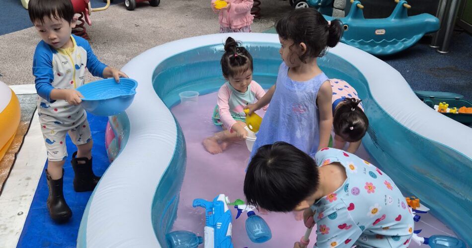 파란색 긴팔 수영복을 입은 지호가 분홍색 물이 담긴 작은 수영장 앞에서 파란색 대야를 들고 서 있다. 수영장 안팎에는 여러 명의 아이들이 물장난감들을 가지고 놀고 있다. 배경에는 어린이집 놀이터 시설들이 보인다.