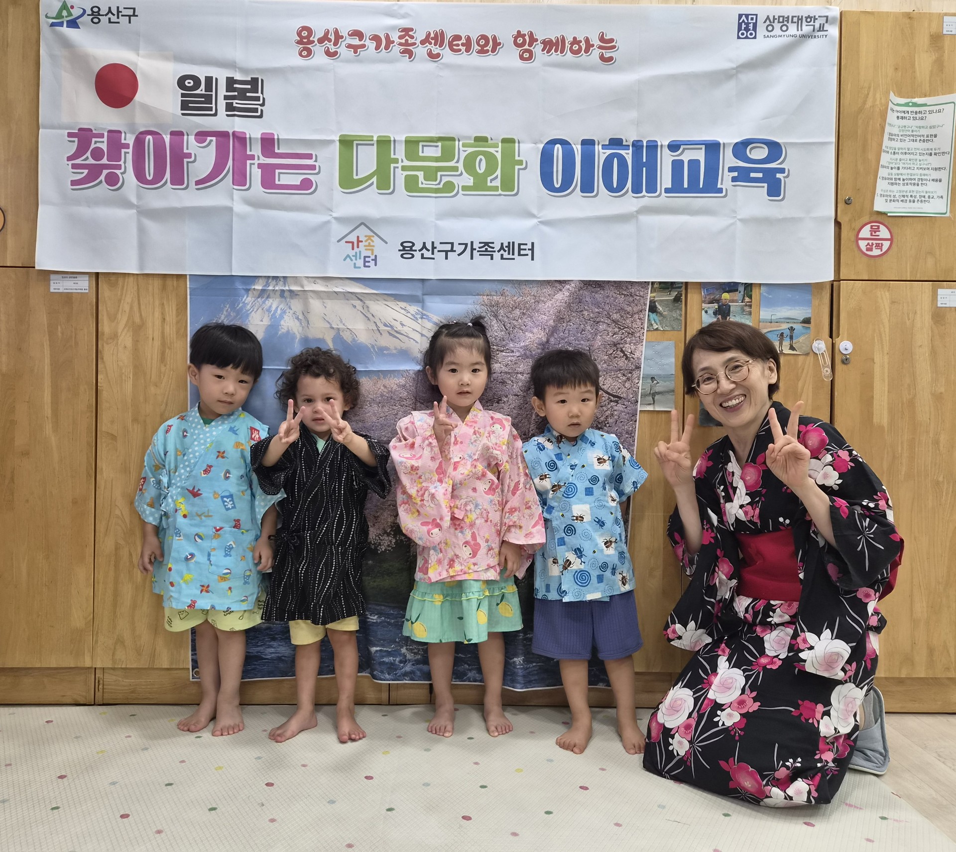 '일본 찾아가는 다문화 이해교육' 배너 앞에서 네 명의 어린아이들이 일본 전통 의상인 유카타를 입고 서 있고, 옆에는 유카타를 입은 여성 선생님이 무릎을 꿇고 V자를 그리며 함께 포즈를 취하고 있다. 지호는 하늘색 유카타를 입고 환하게 웃고 있다.