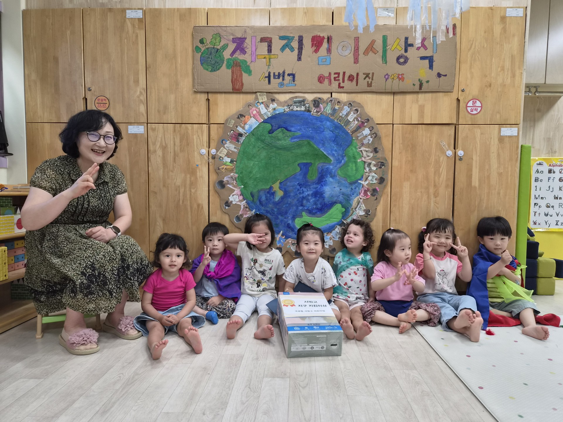 선생님 한 분과 8명의 아이들이 '지구지킴이 사랑어린이집'이라고 적힌 나무 벽과 지구 그림 앞에서 나란히 앉아 밝게 웃으며 단체 사진을 찍고 있다. 가운데에는 작은 책이 놓여있다.