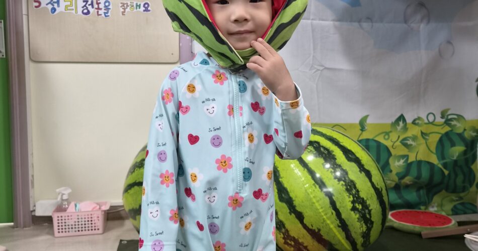 연한 하늘색 꽃무늬 수영복을 입은 어린 지호가 수박 껍질을 연상시키는 초록색과 빨간색 모자를 쓰고 정면을 보고 서 있다. 지호는 살짝 미소 지으며 오른손으로 턱을 가리키고 있고, 배경에는 수박 모양 소품들과 교육 자료가 보인다.
