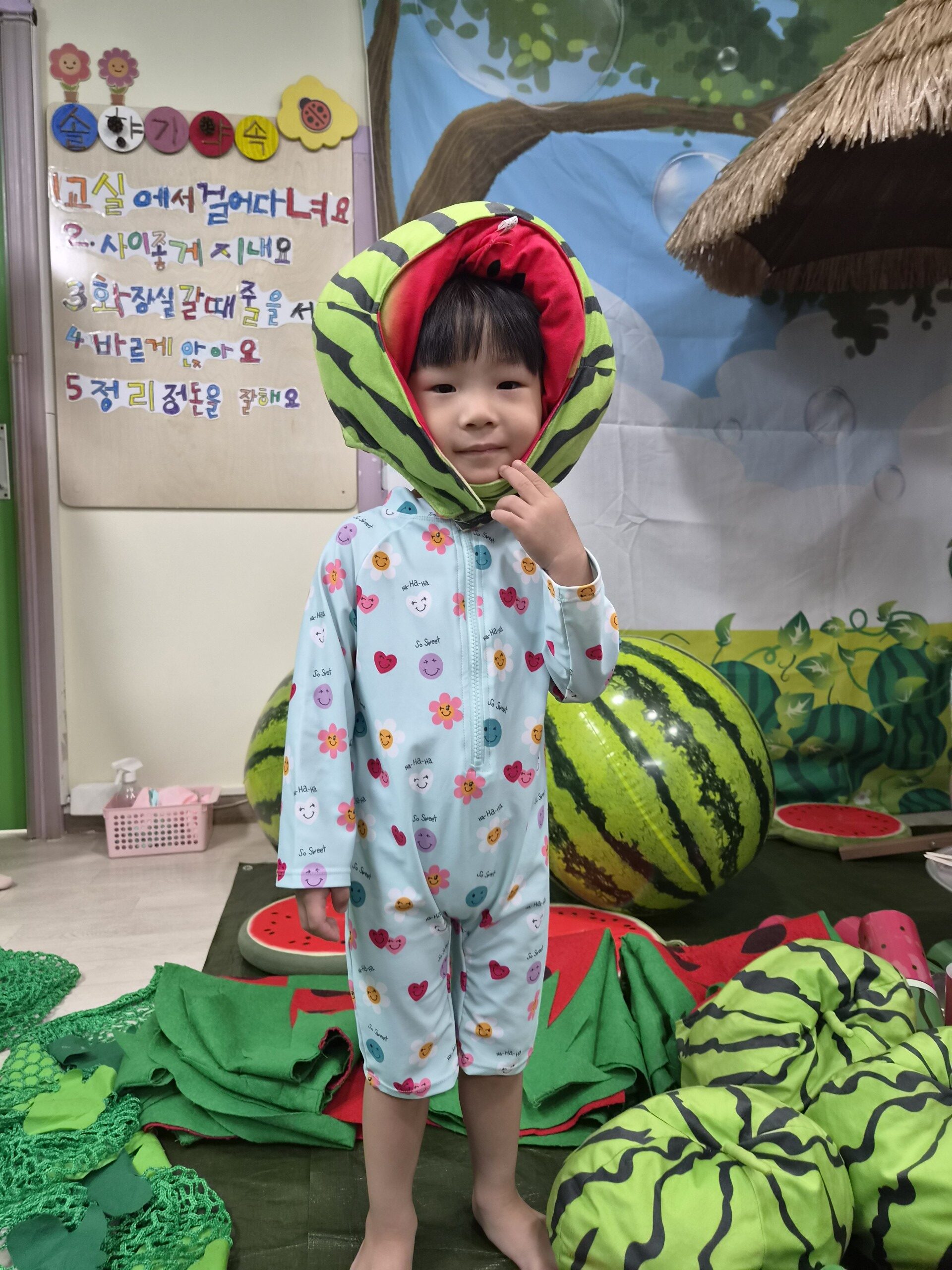 연한 하늘색 꽃무늬 수영복을 입은 어린 지호가 수박 껍질을 연상시키는 초록색과 빨간색 모자를 쓰고 정면을 보고 서 있다. 지호는 살짝 미소 지으며 오른손으로 턱을 가리키고 있고, 배경에는 수박 모양 소품들과 교육 자료가 보인다.
