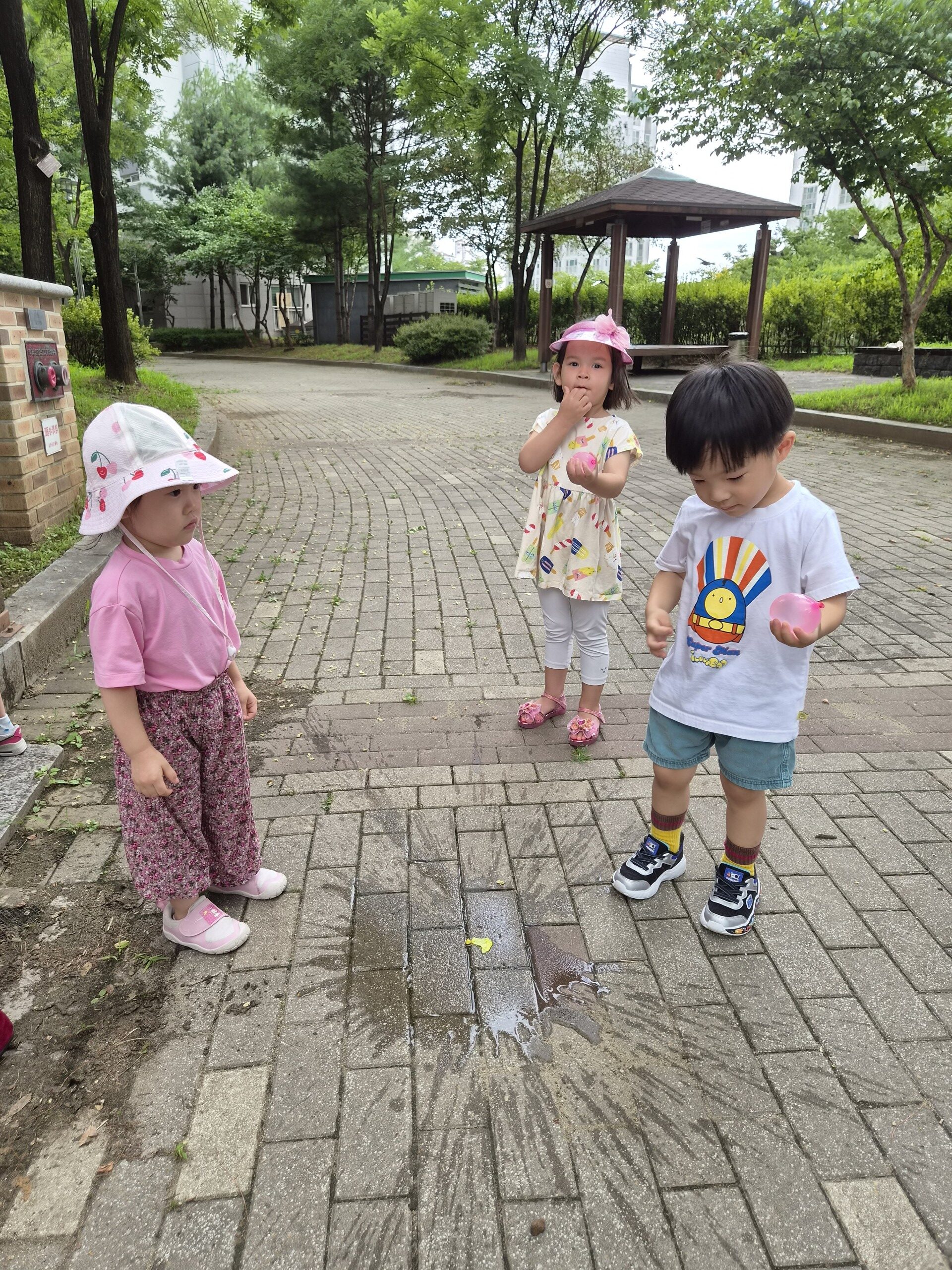 하얀색 기차 그림 티셔츠를 입은 지호와 두 명의 여자아이가 야외 공원 보도블록 위에서 놀고 있다. 지호는 분홍색 물풍선을 들고 바닥에 튀긴 물방울을 바라보고 있다.