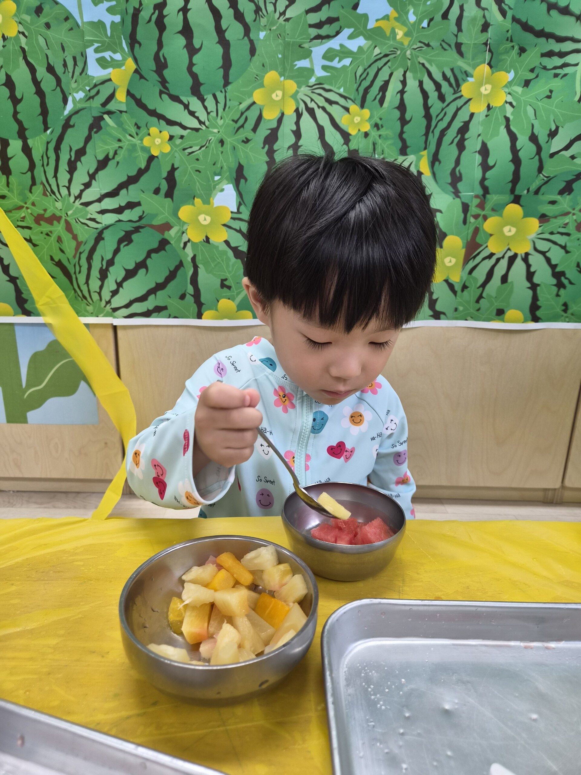 지호가 노란색 테이블에 앉아 작은 스테인리스 그릇에 담긴 깍둑썰기 한 과일을 숟가락으로 떠먹고 있다. 배경은 수박 그림 벽지다.