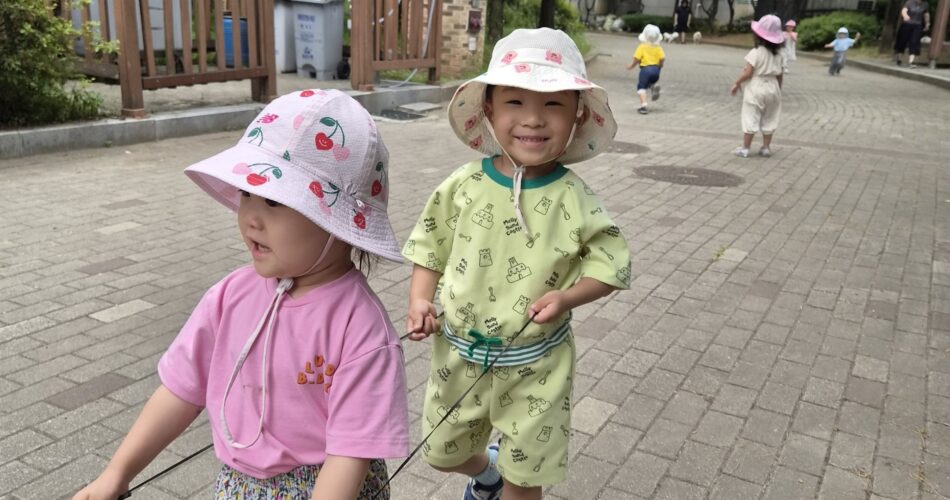 초록색 옷과 체리 무늬 모자를 쓴 지호가 분홍색 옷을 입은 여자아이와 나란히 서서 줄을 잡고 밝게 웃으며 걷고 있다. 주변에는 다른 아이들도 함께 산책 중이며, 배경은 보도블록이 깔린 길이다.