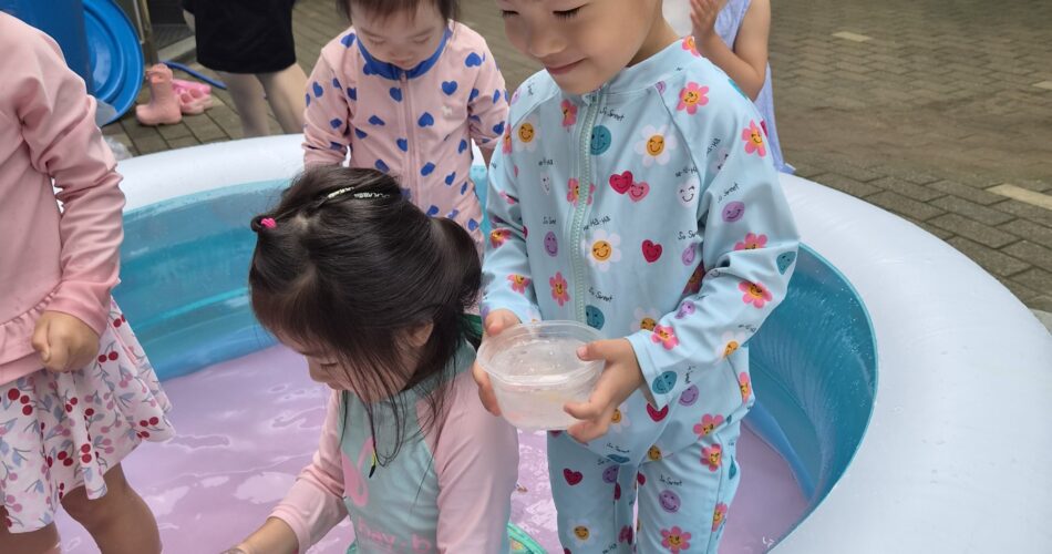 파란색 물놀이 수영복을 입은 지호가 야외 간이 수영장에서 친구들과 함께 물장구를 치며 컵에 물을 담고 있는 모습. 다른 친구들도 수영복을 입고 물놀이를 즐기고 있으며, 뒤편에는 빨간 티셔츠를 입은 어른의 모습이 보인다.